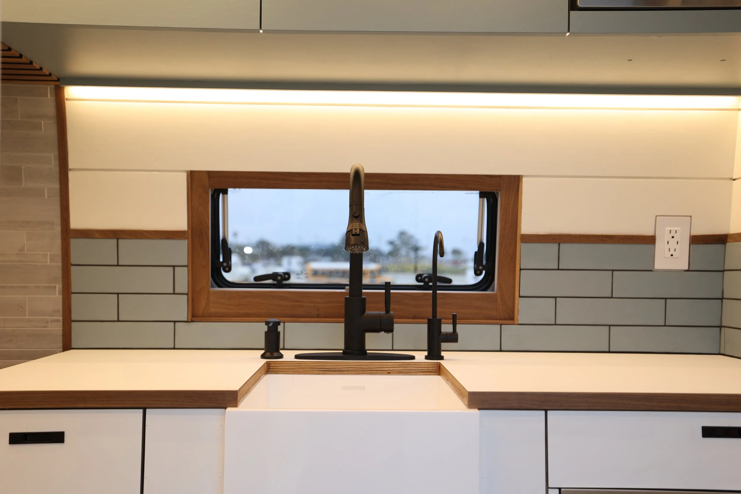 Modern kitchen sink with black faucet and soap dispenser beneath a window with wooden frame, gray subway tile backsplash, and white cabinets.
