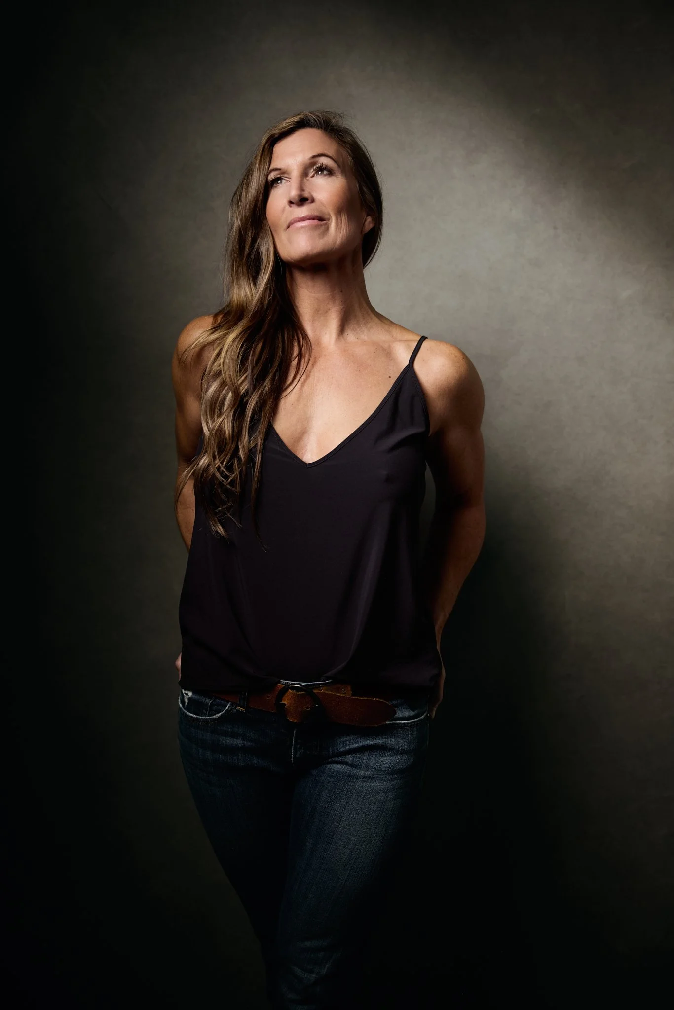 A woman with long wavy brown hair, wearing a black camisole top and blue jeans, standing against a gradient background with a thoughtful expression.