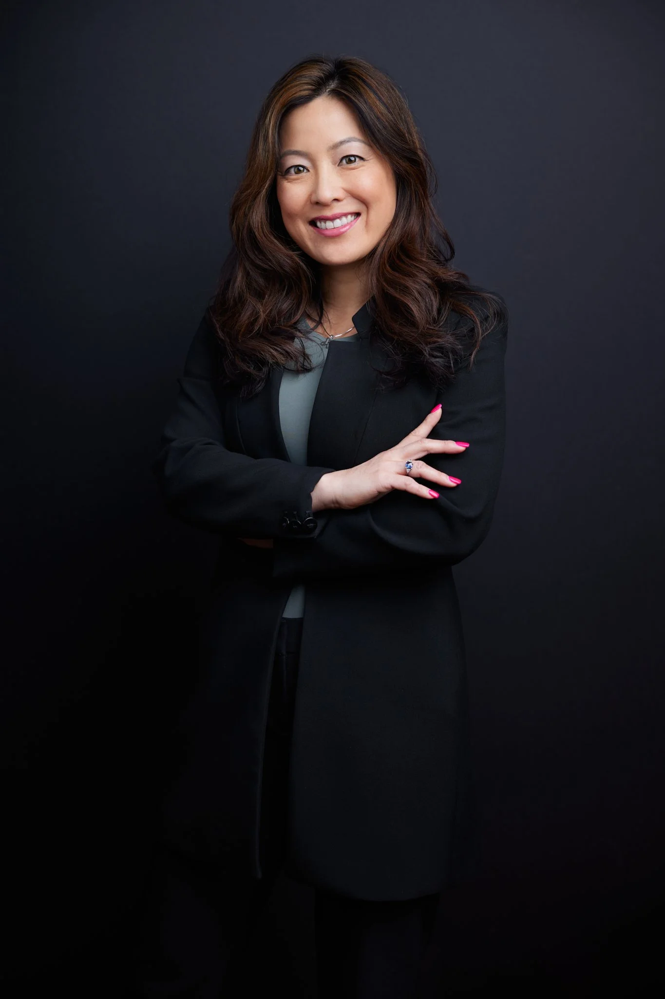 A woman with long, wavy hair, smiling and wearing a black blazer and gray top, standing against a dark background.
