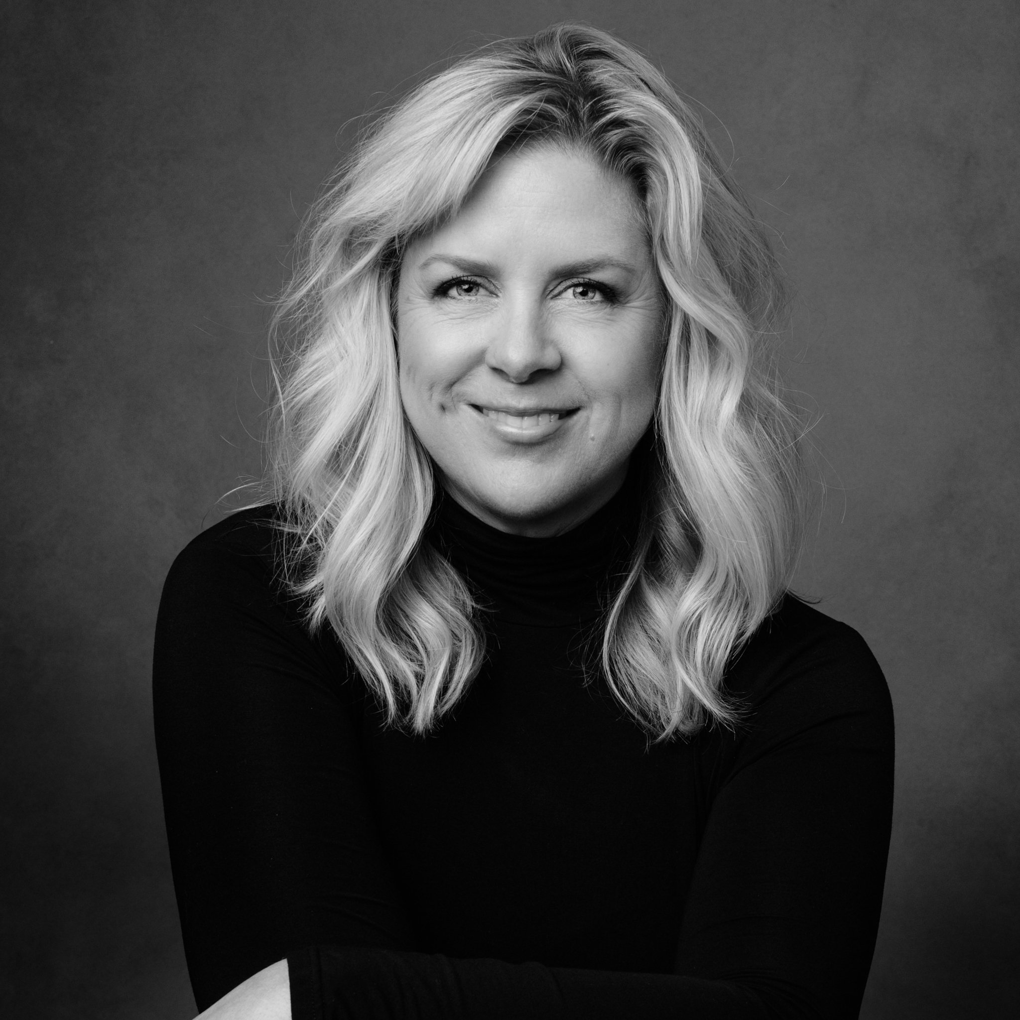 Black and white portrait of a woman with wavy blonde hair wearing a black turtleneck, smiling at the camera.