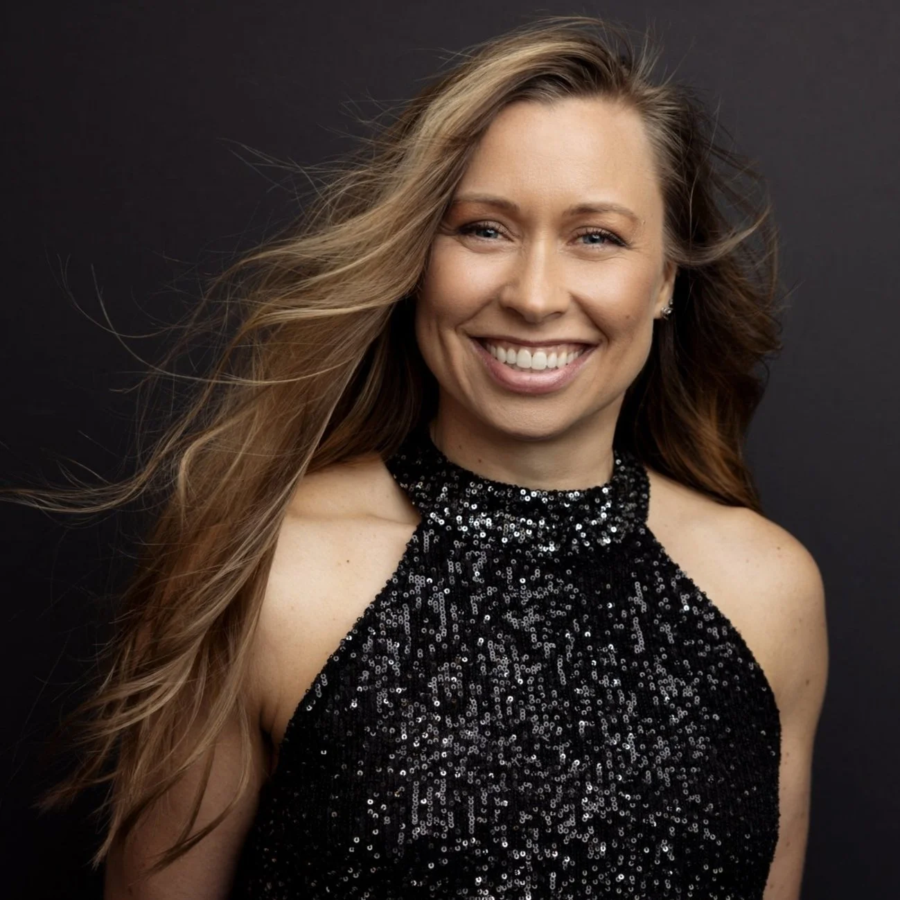 A woman with long, wavy blonde hair, smiling, wearing a black sequined halter dress, against a dark background.