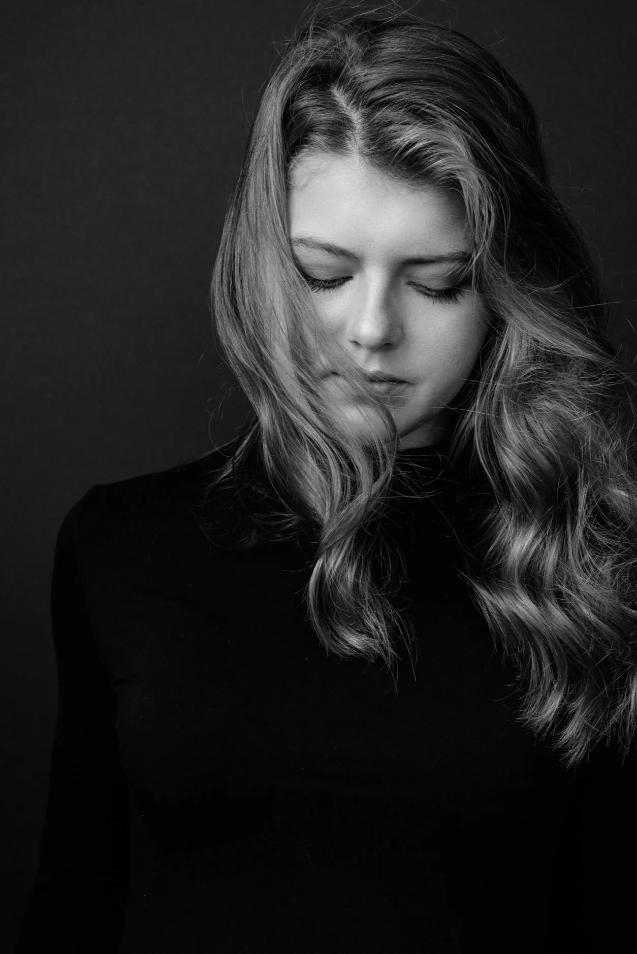 A black and white portrait of a young woman with wavy hair, her eyes closed, wearing a dark top, against a plain background.