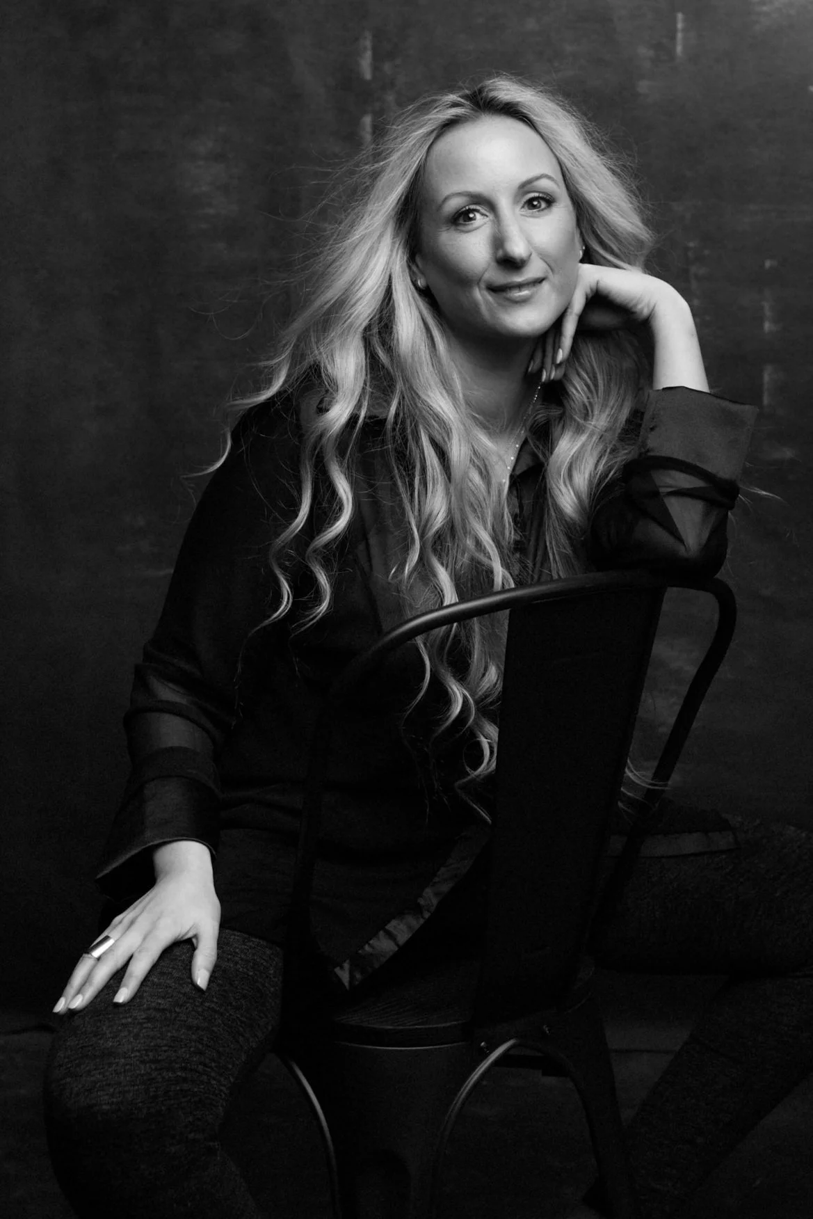 A black and white portrait of a woman with long, wavy hair, sitting on a chair, smiling, with her hand resting on her chin against a dark background.