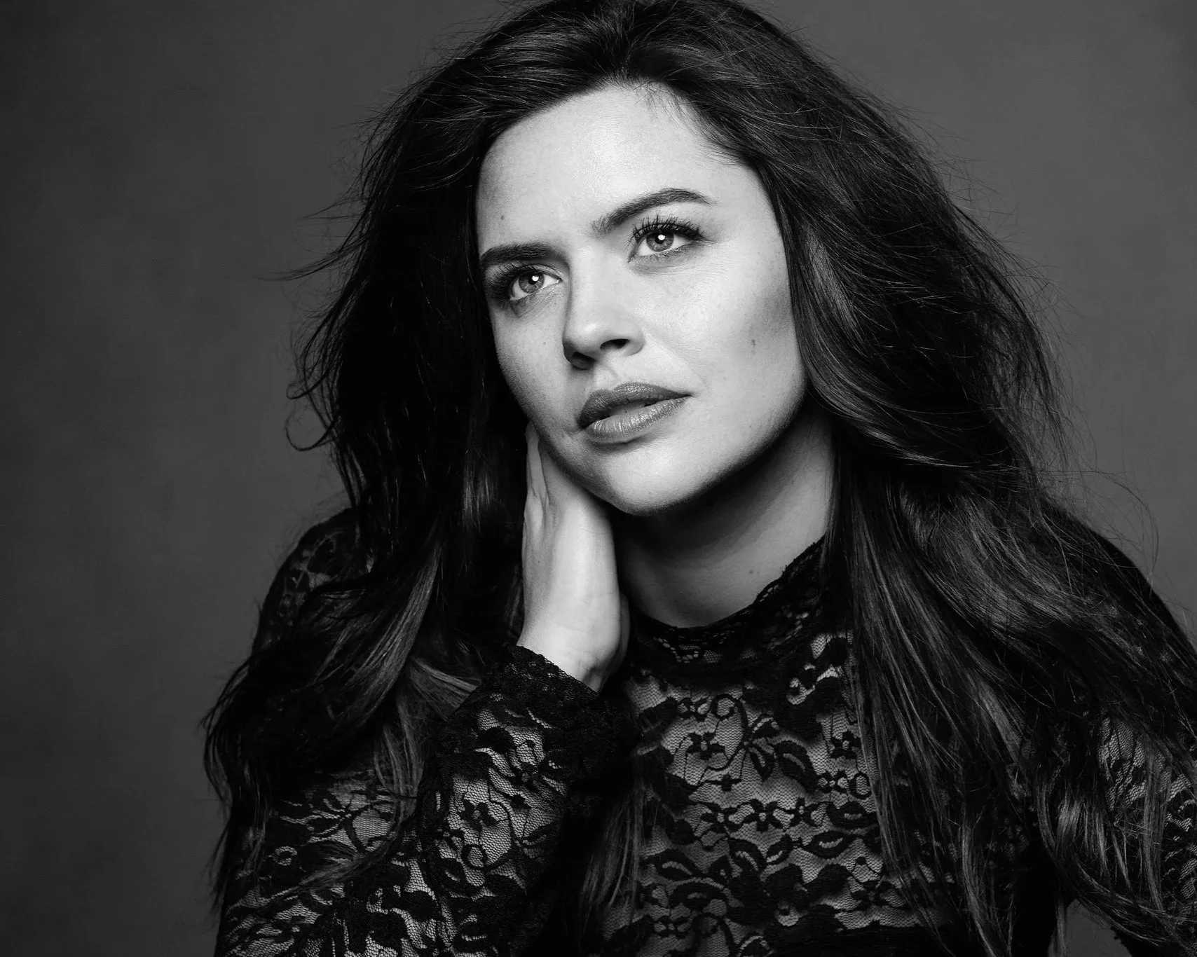 Black and white portrait of a woman with long wavy hair wearing a lace top, resting her face on her hand.