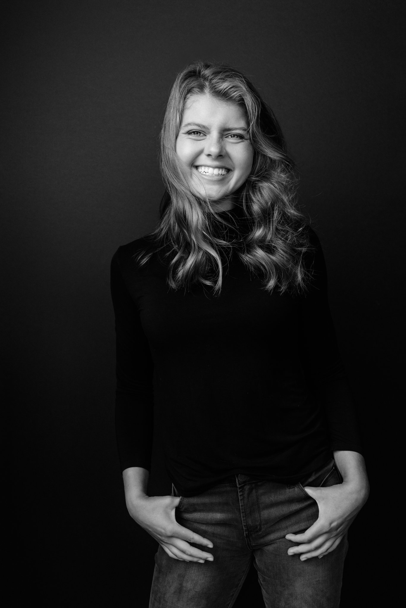 Black and white portrait of a woman with wavy hair, smiling, wearing a long-sleeve top and jeans, standing against a dark background.