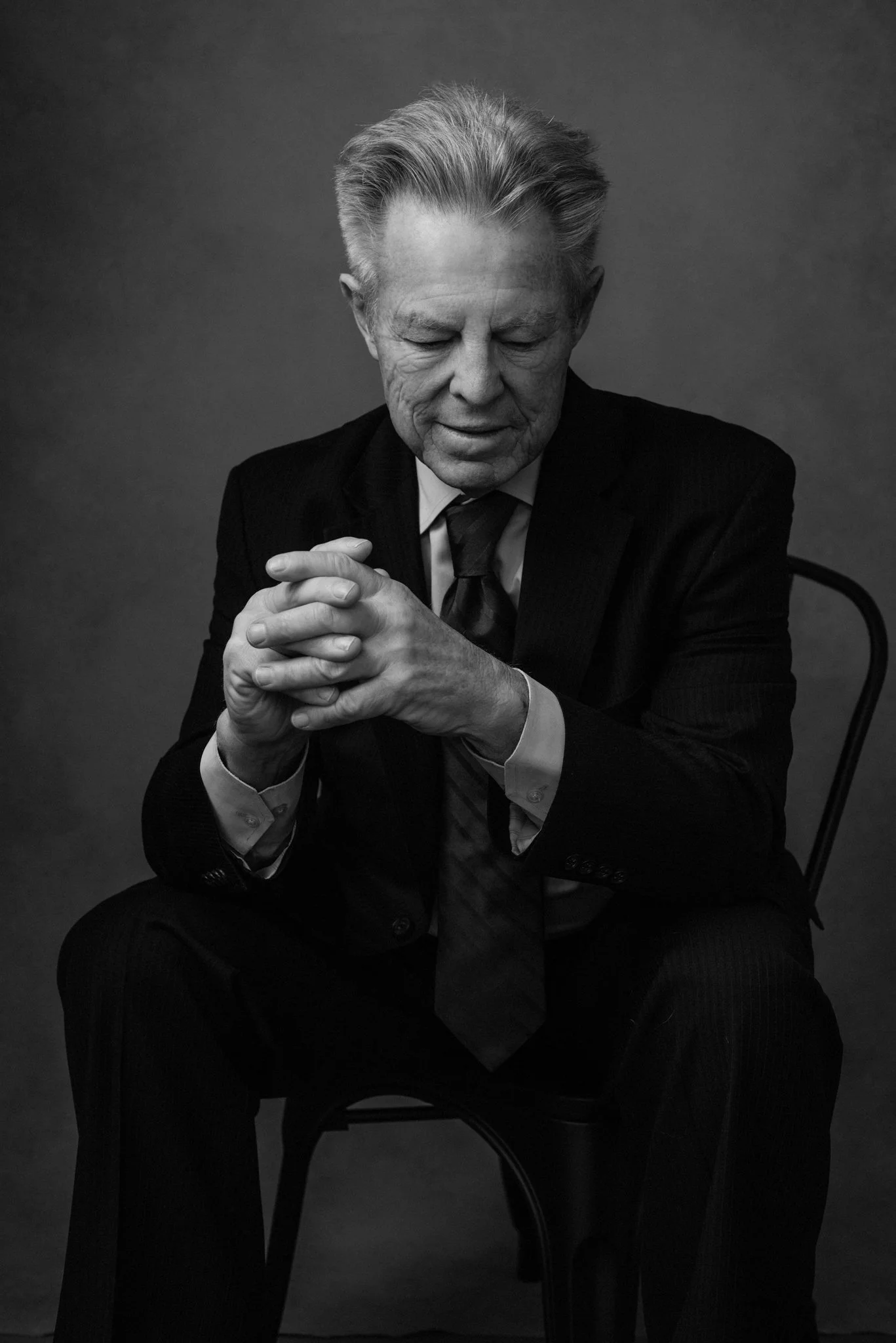 A black and white photo of an elderly man in a suit sitting on a chair with his eyes closed and hands clasped in front of him.