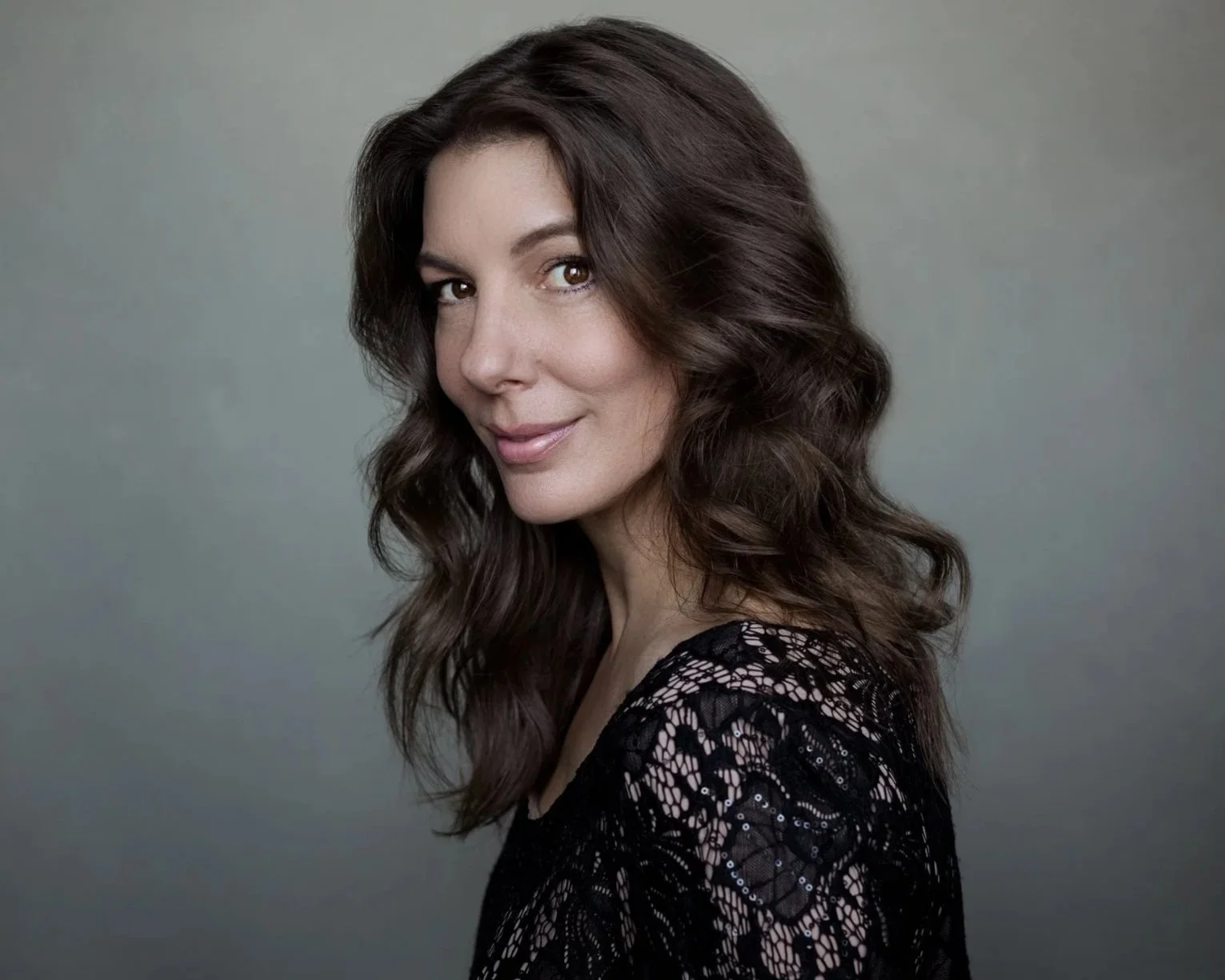 A woman with long, wavy brown hair wearing a black lace top, smiling softly at the camera against a plain background.