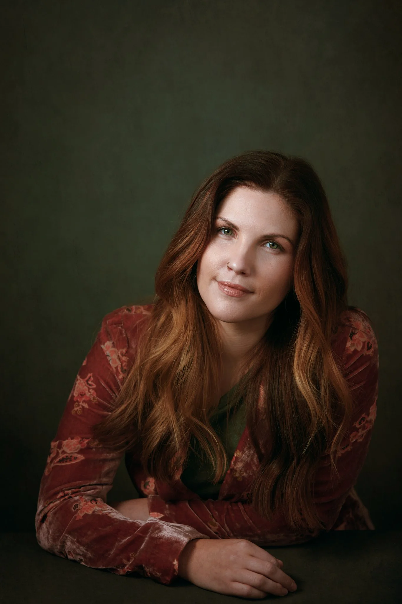A young woman with long wavy brown hair, wearing a patterned velvet red blazer, posing against a dark background.