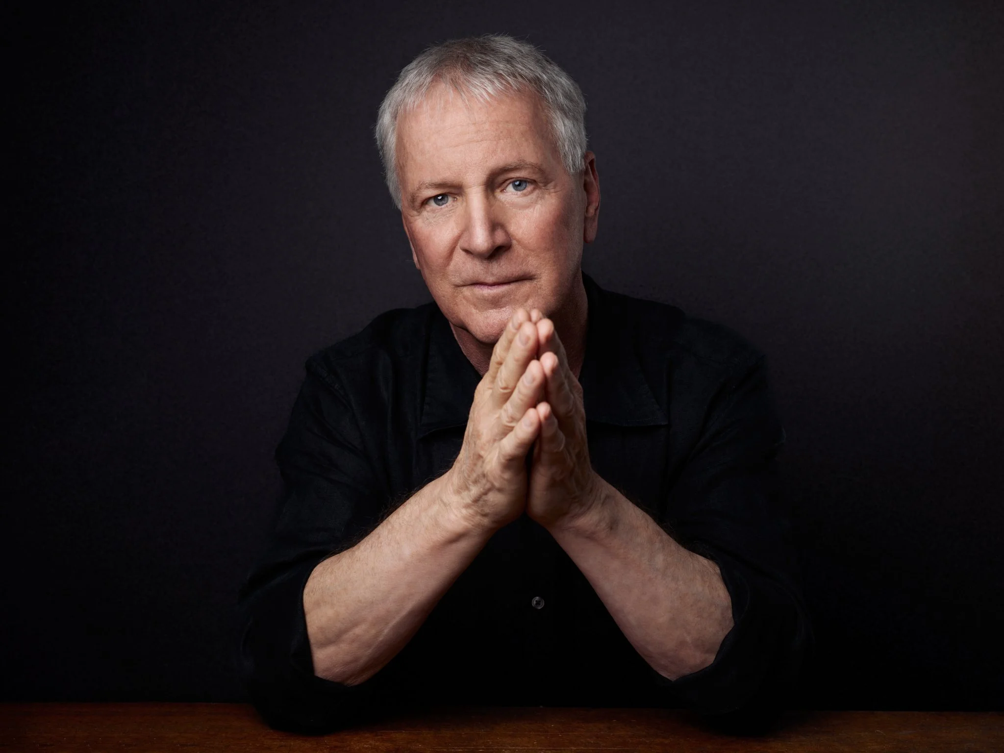 Older man with gray hair and blue eyes wearing a black shirt, pressing his hands together near his face, against a dark background.
