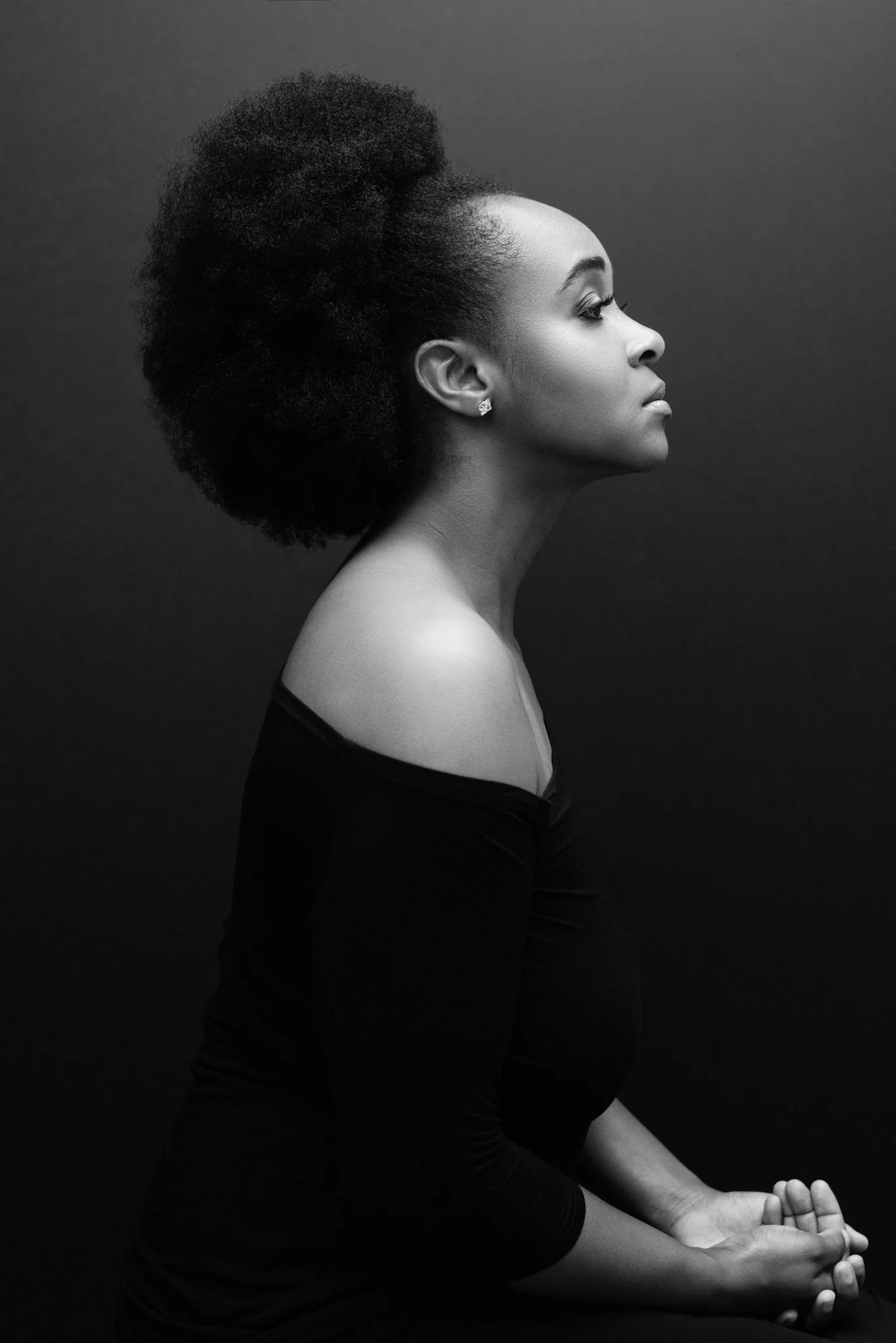 A black-and-white photo of a woman with curly afro hair sitting in a meditative pose against a dark background. She is wearing a long-sleeve off-shoulder top and earring, with her eyes closed and hands clasped.