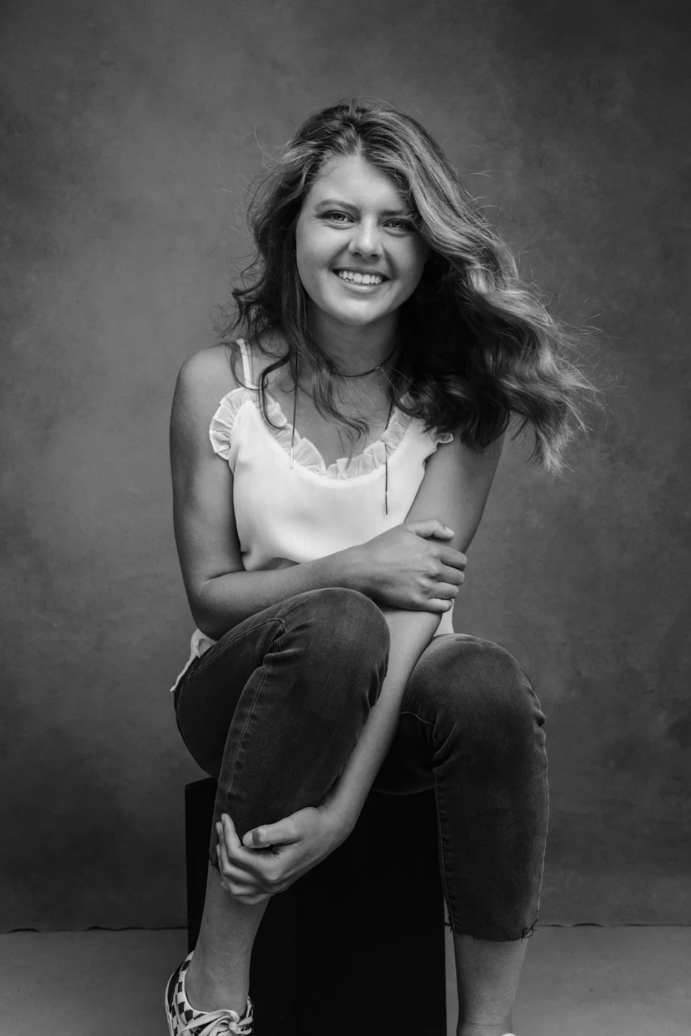 Black and white portrait of a smiling young woman with wavy hair, sitting on a stool against a plain background, wearing a white sleeveless top and dark jeans.
