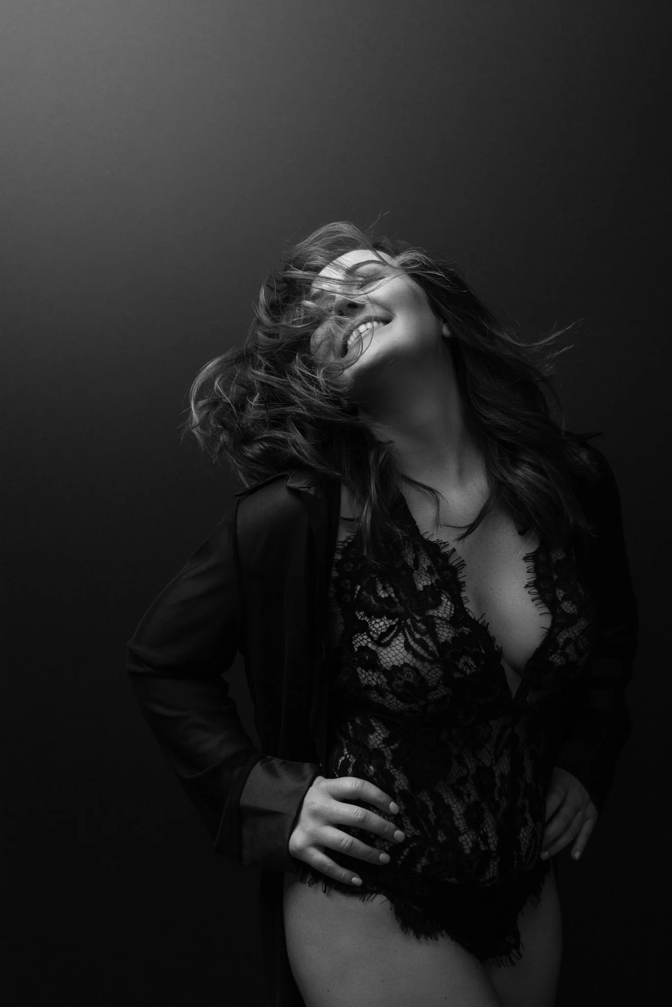 A woman smiling, with her hair blowing, wearing a lace top and a dark jacket, standing against a dark background.