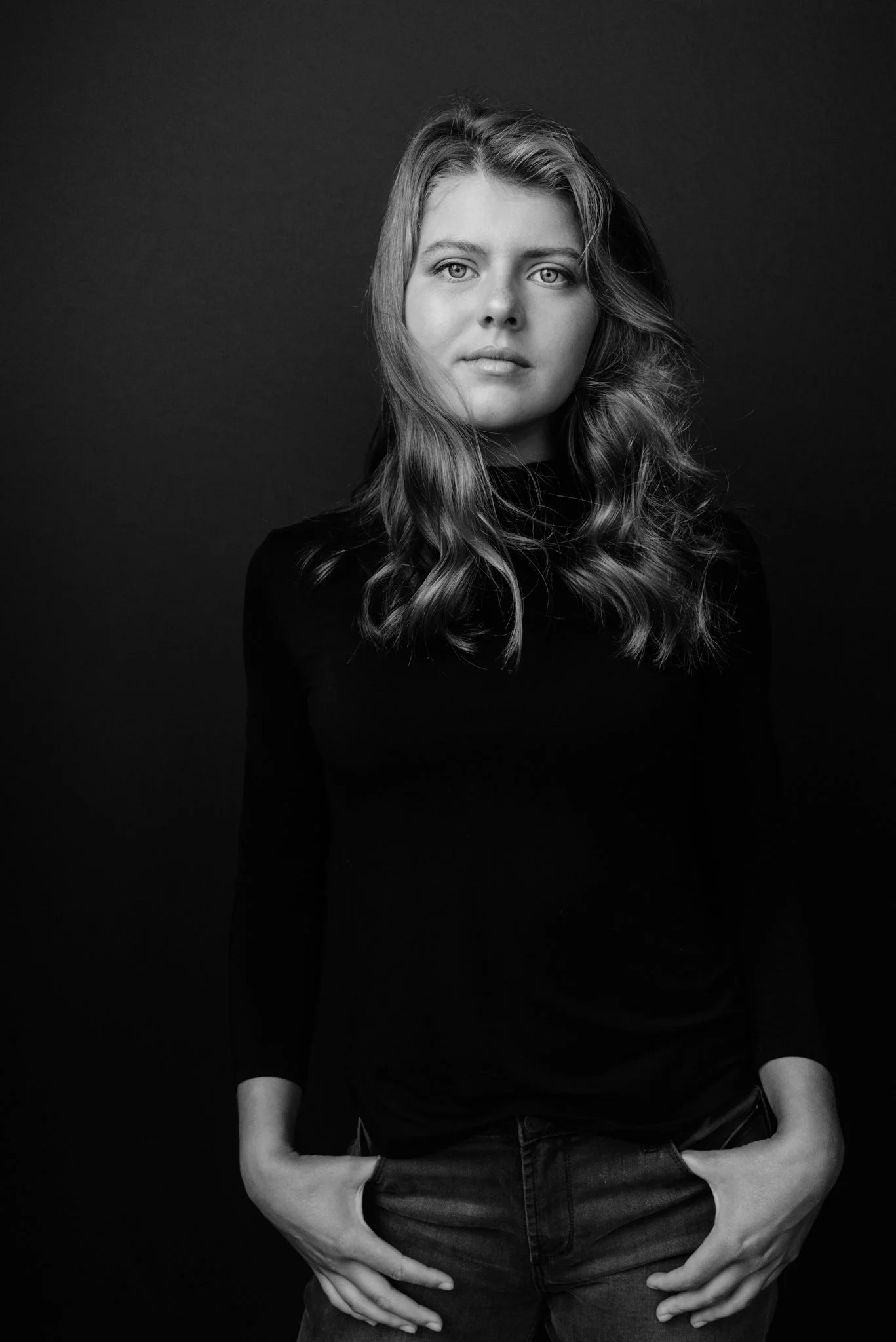 Black and white portrait of a young woman with wavy hair, wearing a long-sleeved top, standing against a dark background with her hands in her pockets, looking directly at the camera.