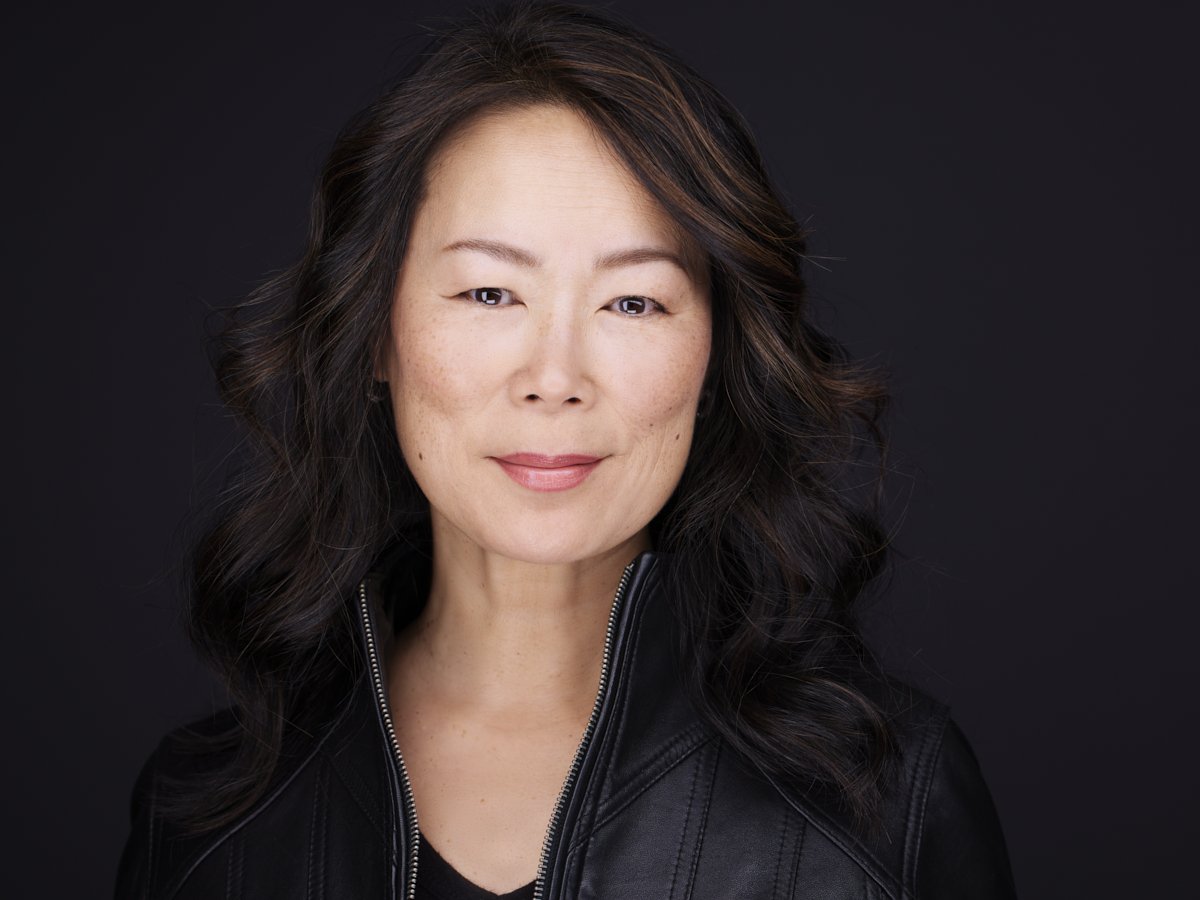 A portrait of a woman with long wavy dark hair, light makeup, and a subtle smile, wearing a black leather jacket against a black background.