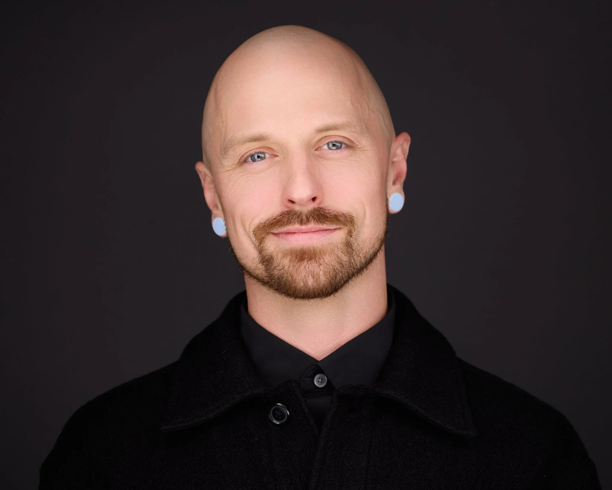 A man with a shaved head, light beard, and light blue eyes, wearing a black coat and shirt, smiling against a dark background.