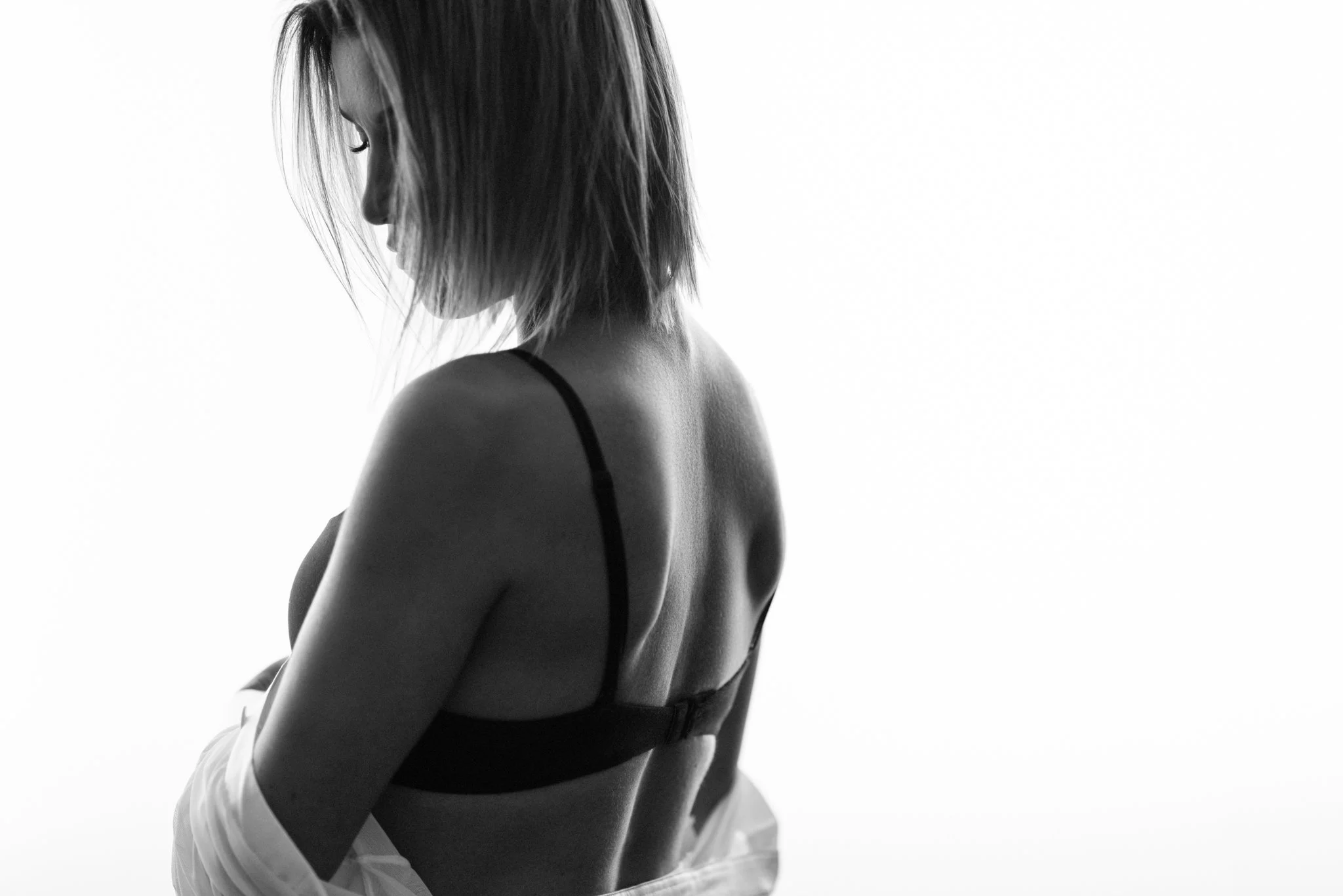 Black and white photo of a woman with shoulder-length hair wearing a bra, looking down with her face turned to the side.