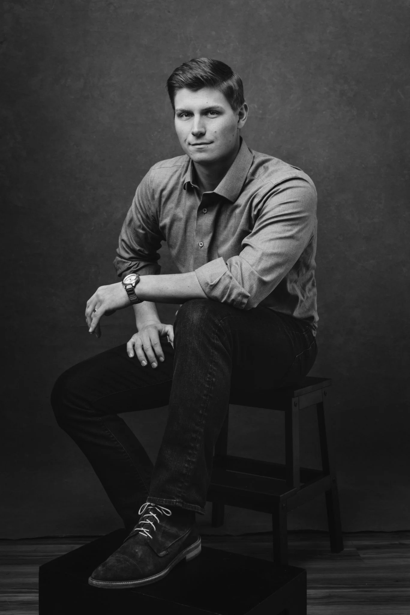 A young man with dark hair and a slight smile, wearing a long-sleeve button-up shirt, dark jeans, and dress shoes, sitting on a stool against a dark textured background.