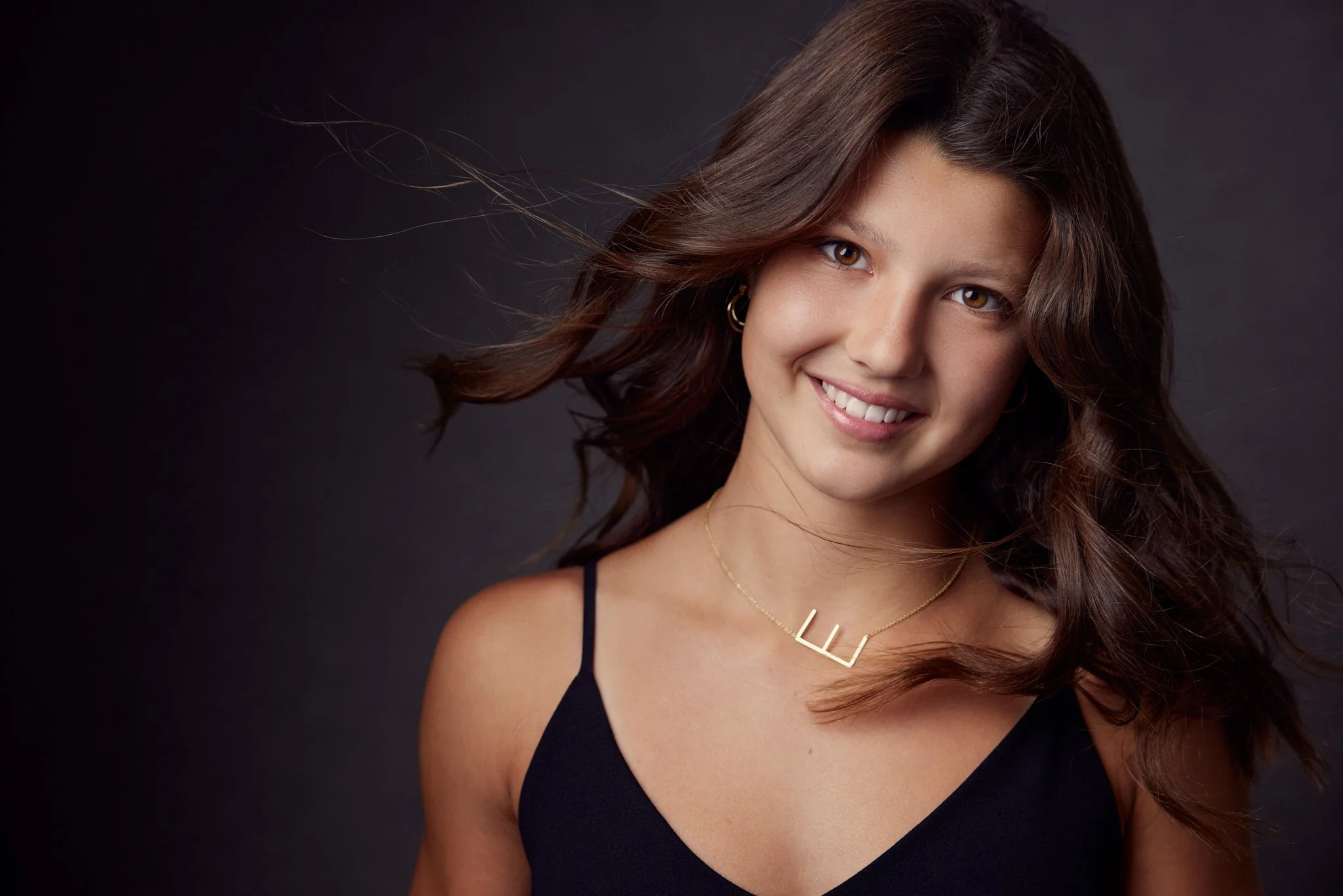 A young woman with shoulder-length wavy brown hair, wearing a black spaghetti strap top, gold hoop earrings, and a gold necklace with the letter 'L', smiling against a dark background.