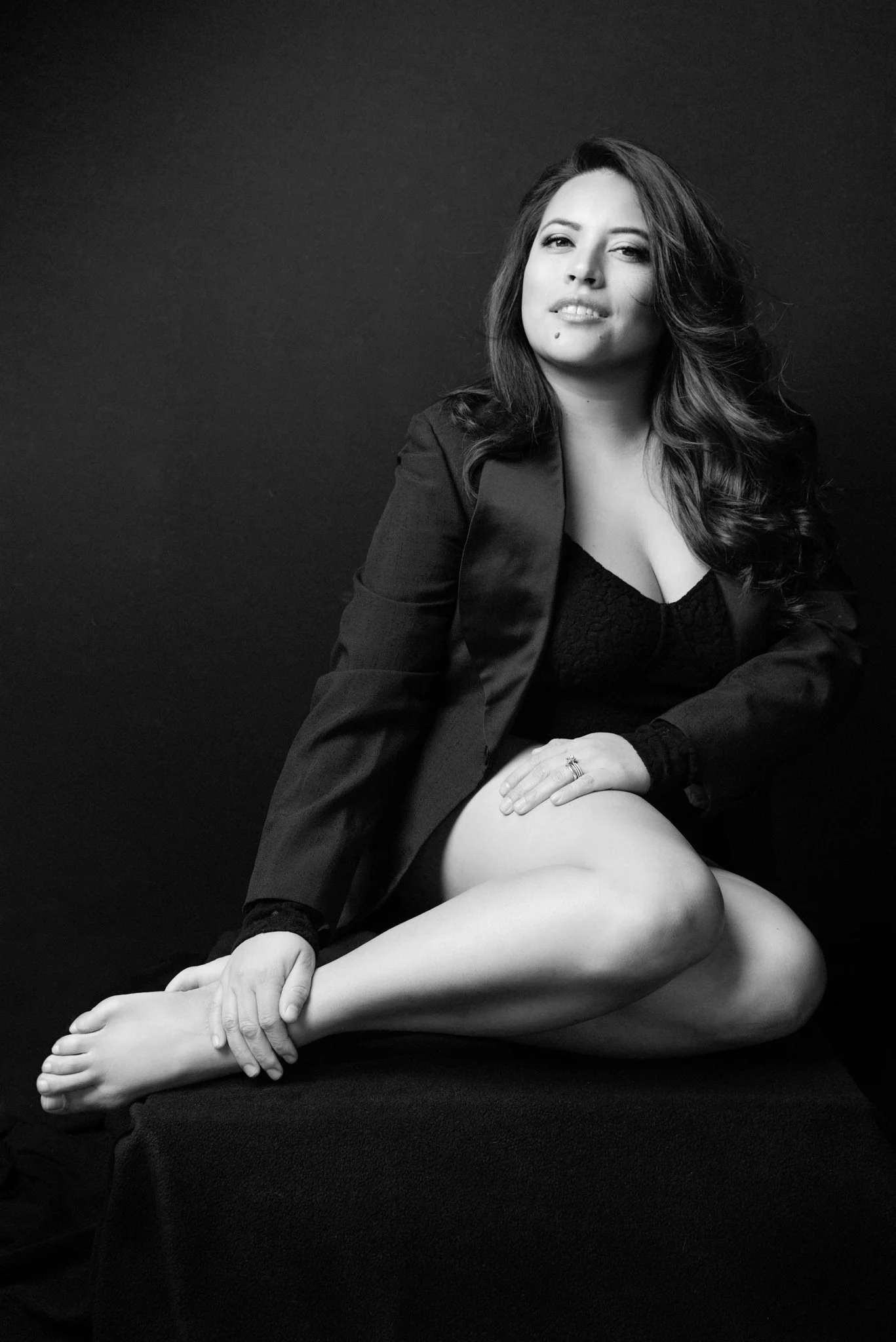 Black and white portrait of a woman with long, wavy hair, sitting with one leg crossed over the other, dressed in a dark blazer and lace top, against a plain dark background.