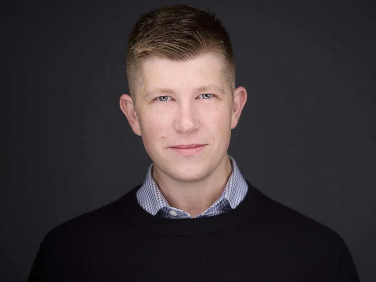 A young man with short light brown hair and blue eyes, wearing a black sweater over a checkered collared shirt, against a dark background.