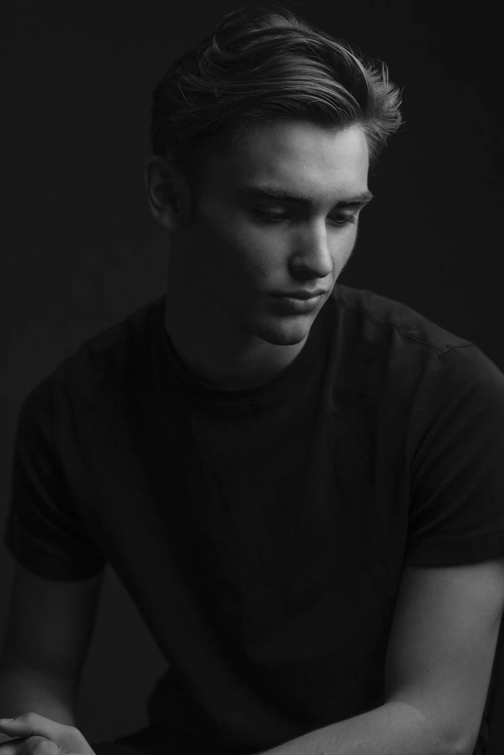 A black and white photograph of a young man with styled hair, looking down with a contemplative expression, against a dark background.