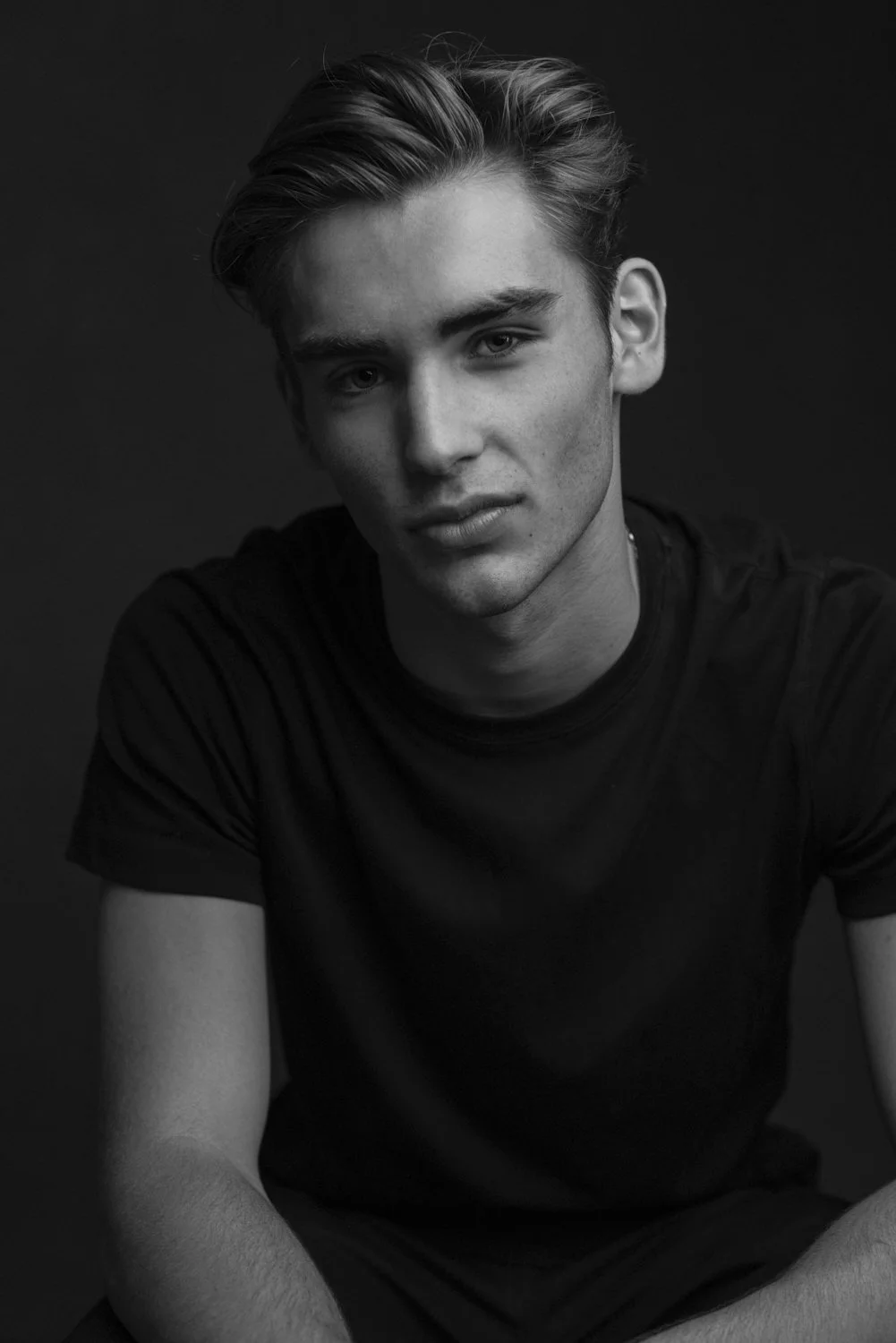 Black and white portrait of a young man with styled hair wearing a black t-shirt, against a dark background.