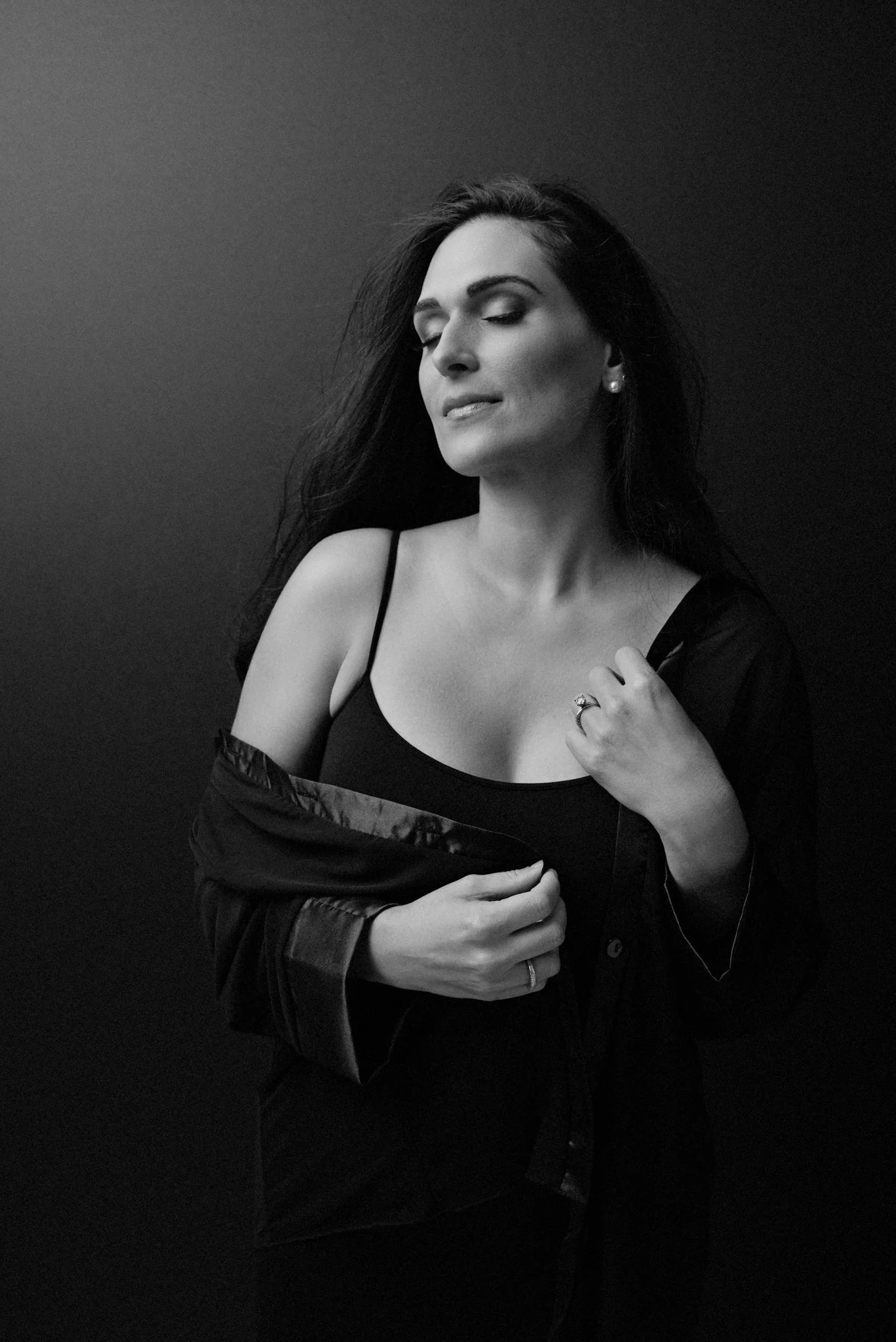 A woman with dark hair, wearing a black tank top and a partially open black shirt, standing against a dark background with her eyes closed.