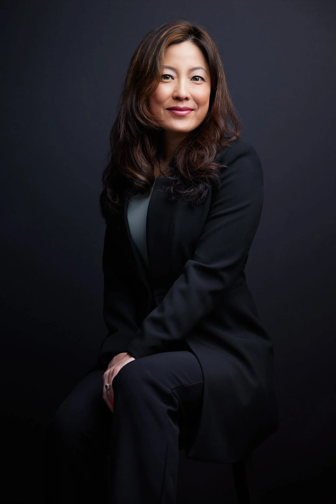 A woman with wavy brown hair smiling, wearing a black blazer and black pants against a dark background.
