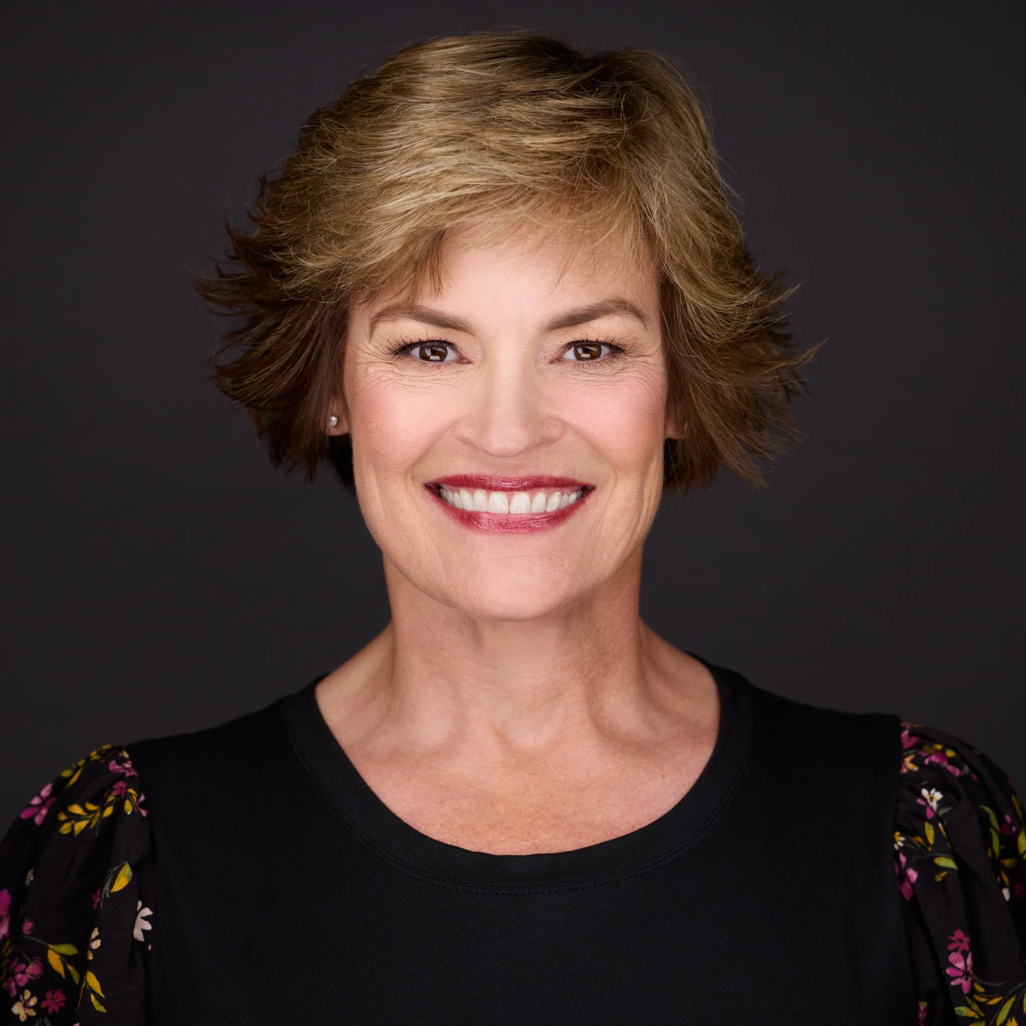 Close-up of a smiling middle-aged woman with short, light brown hair and brown eyes, wearing a black top with floral sleeves against a dark background.