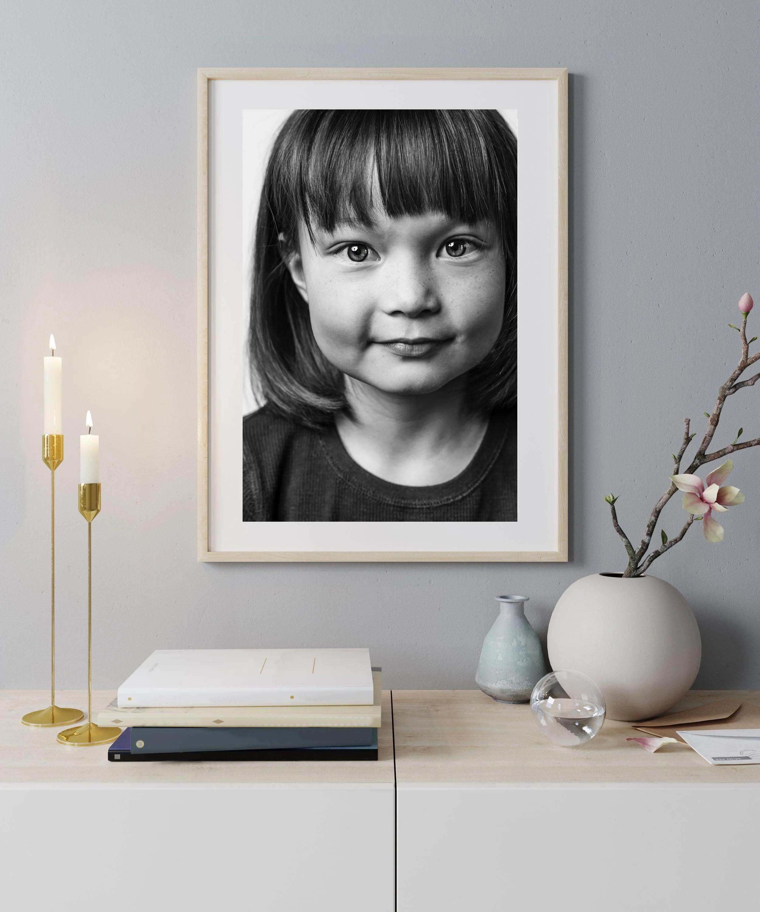 A black and white portrait of a young girl with straight hair and bangs, framed in a light-colored wooden frame, hanging on a wall.
