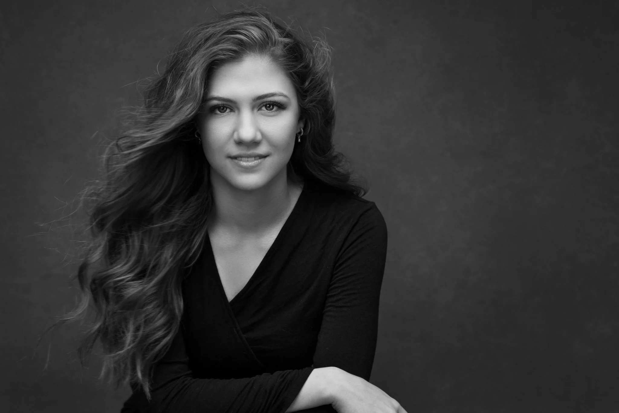 Black and white portrait of a young woman with long, wavy hair, wearing a black top, smiling slightly, against a dark background.