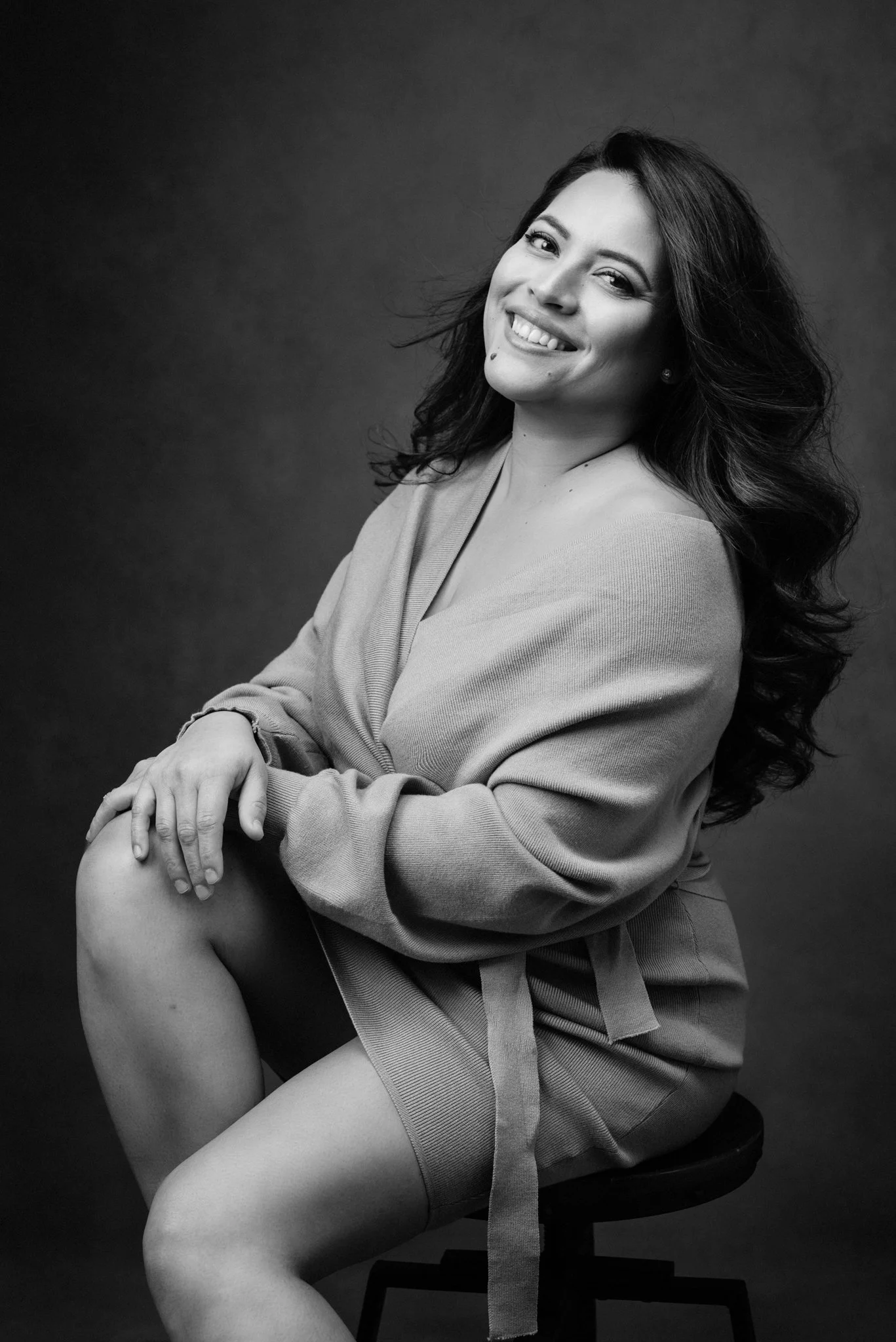 Black and white portrait of a smiling woman with long wavy hair, sitting on a stool, wearing a jacket and shorts, against a dark background.