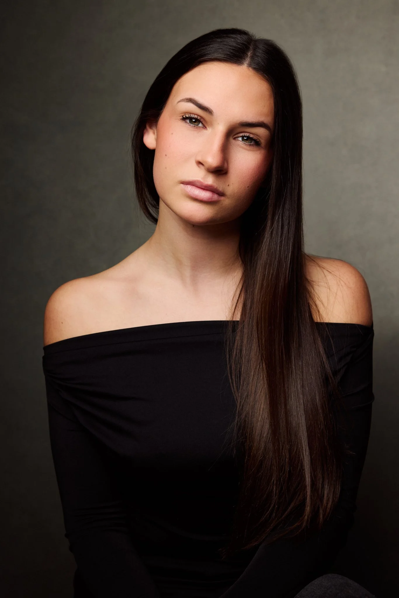 A young woman with long straight dark hair, wearing an off-the-shoulder black top, looking directly at the camera with a neutral expression.