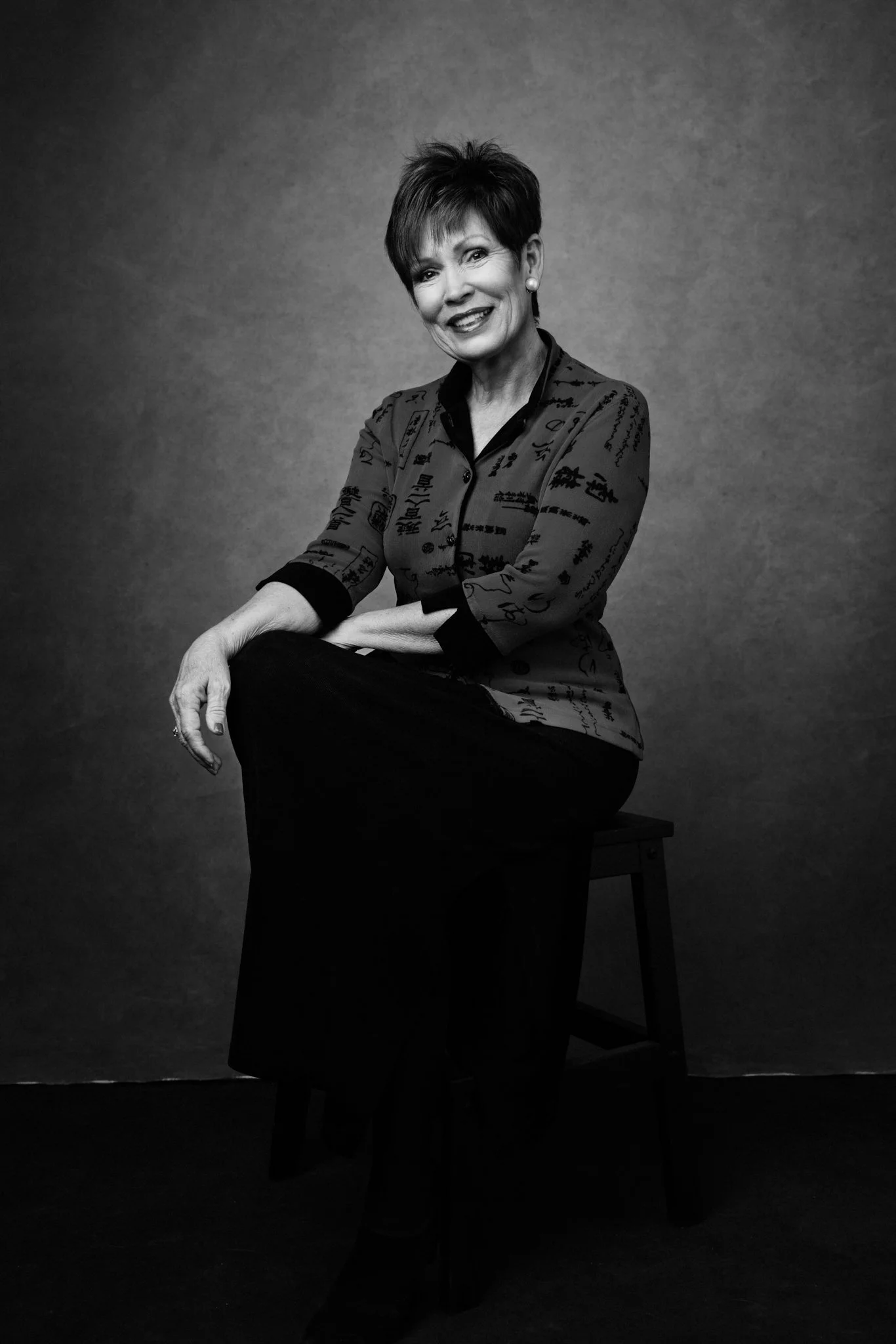 Black and white portrait of a smiling middle-aged woman sitting on a stool against a plain background, wearing a patterned blouse and dark pants.
