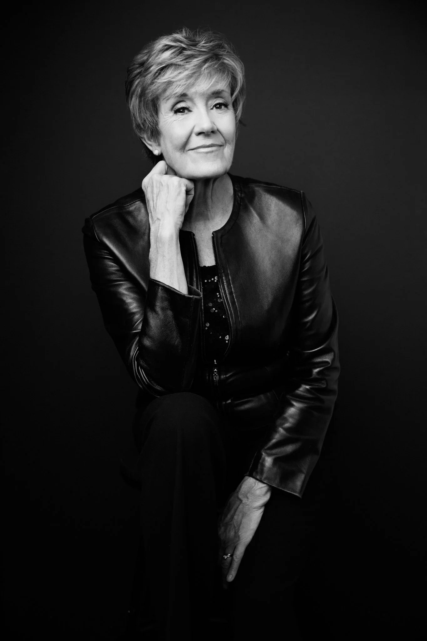 Black and white portrait of an older woman with short hair, wearing a leather jacket, smiling softly, with one hand resting on her chin.
