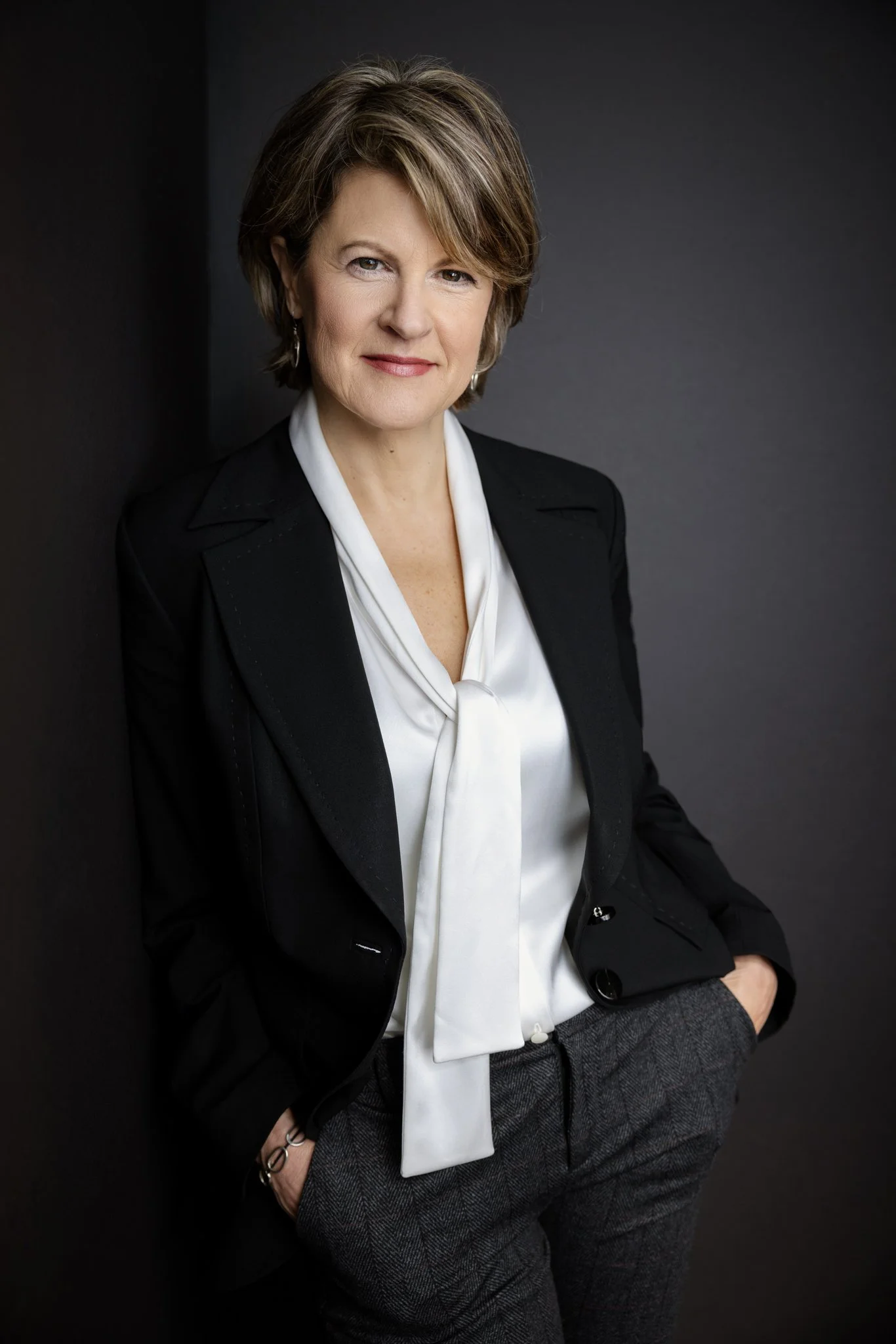 A professional woman with short brown hair, wearing a black blazer over a white blouse, sitting against a dark background.