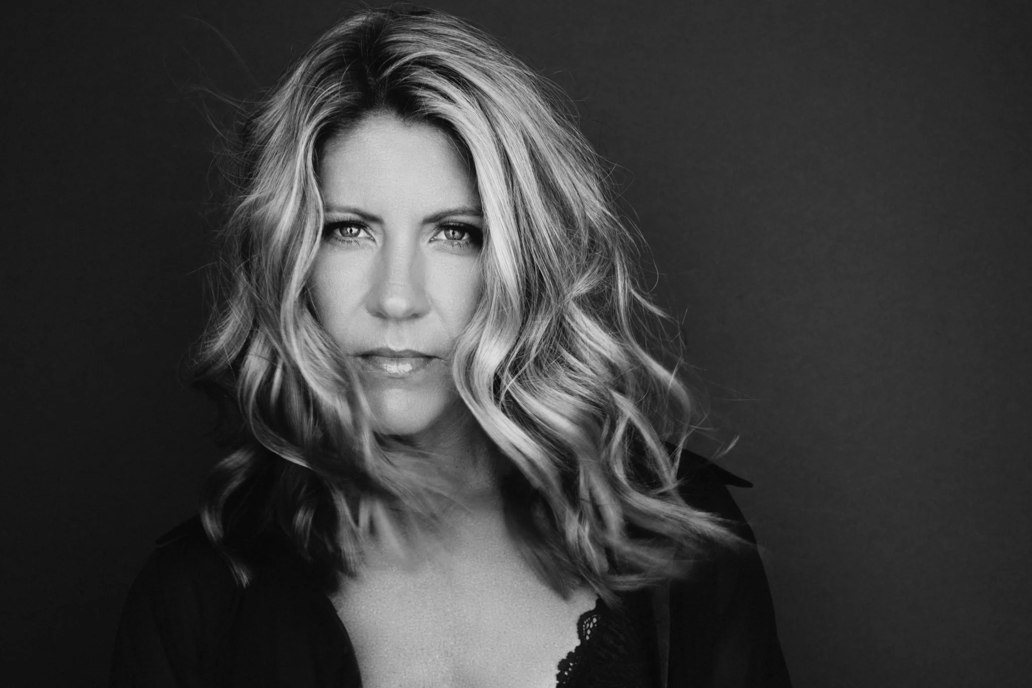 Black and white portrait of a woman with wavy blonde hair, looking directly at the camera, against a plain dark background.