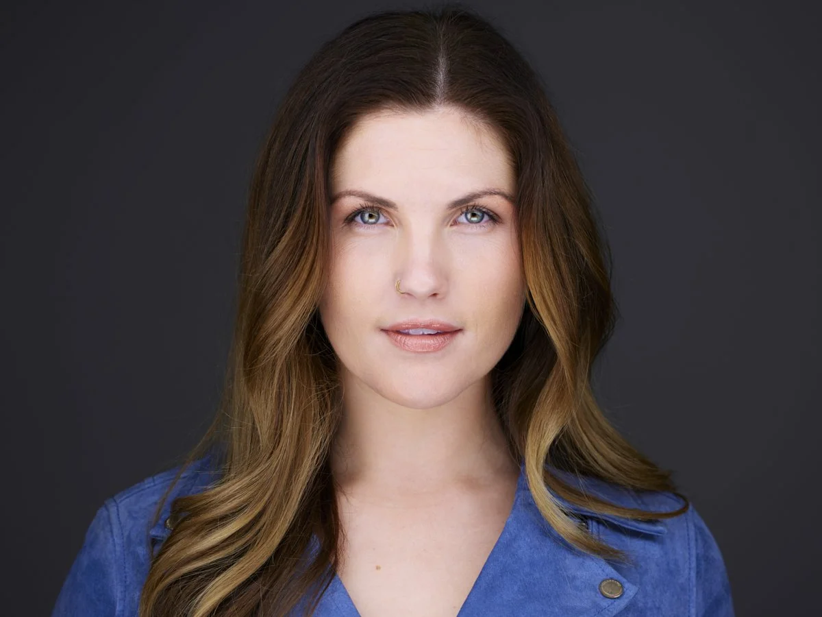 Portrait of a young woman with long wavy brown hair, blue eyes, light skin, wearing a blue top, against a dark background.