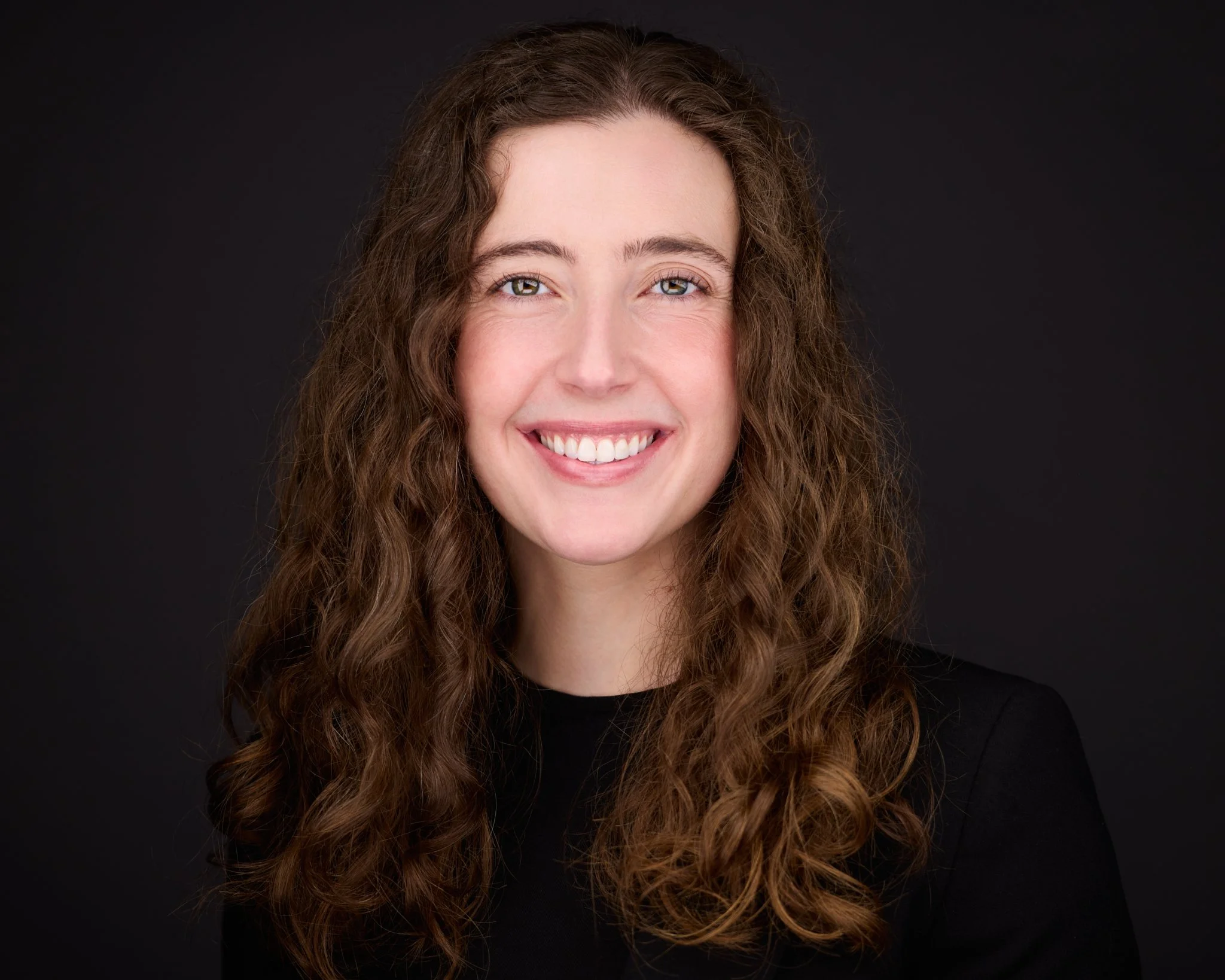 A young woman with long, curly brown hair smiling at the camera against a black background.