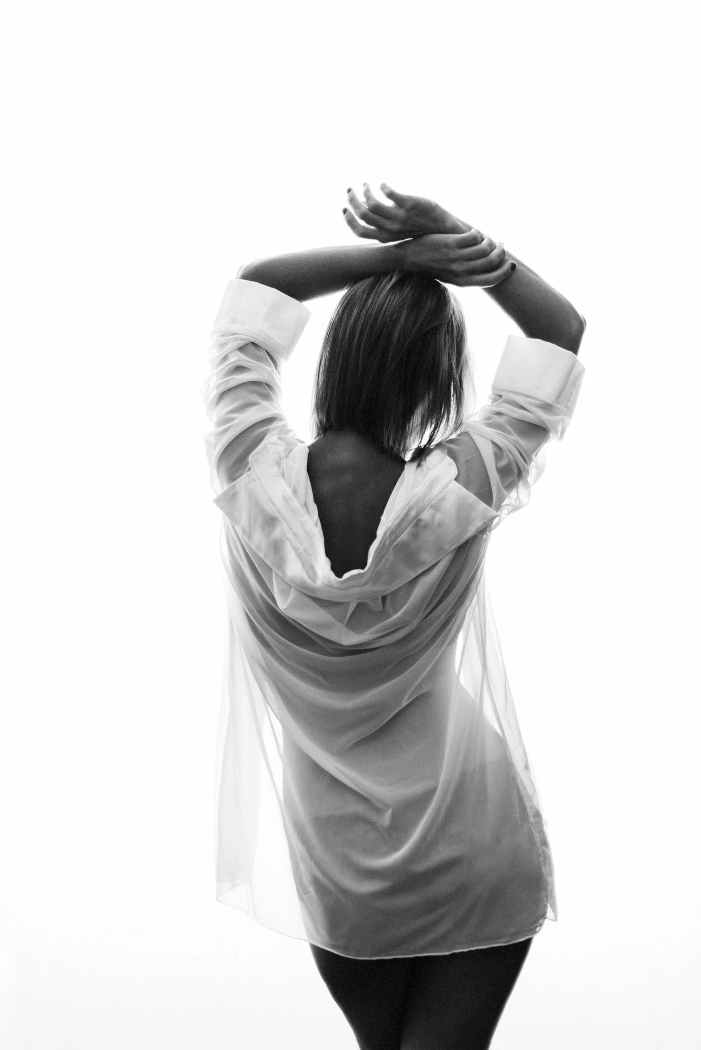 Black and white photo of a woman with shoulder-length hair, wearing a light-colored open shirt and dark pants, with her arms raised above her head and hands touching.