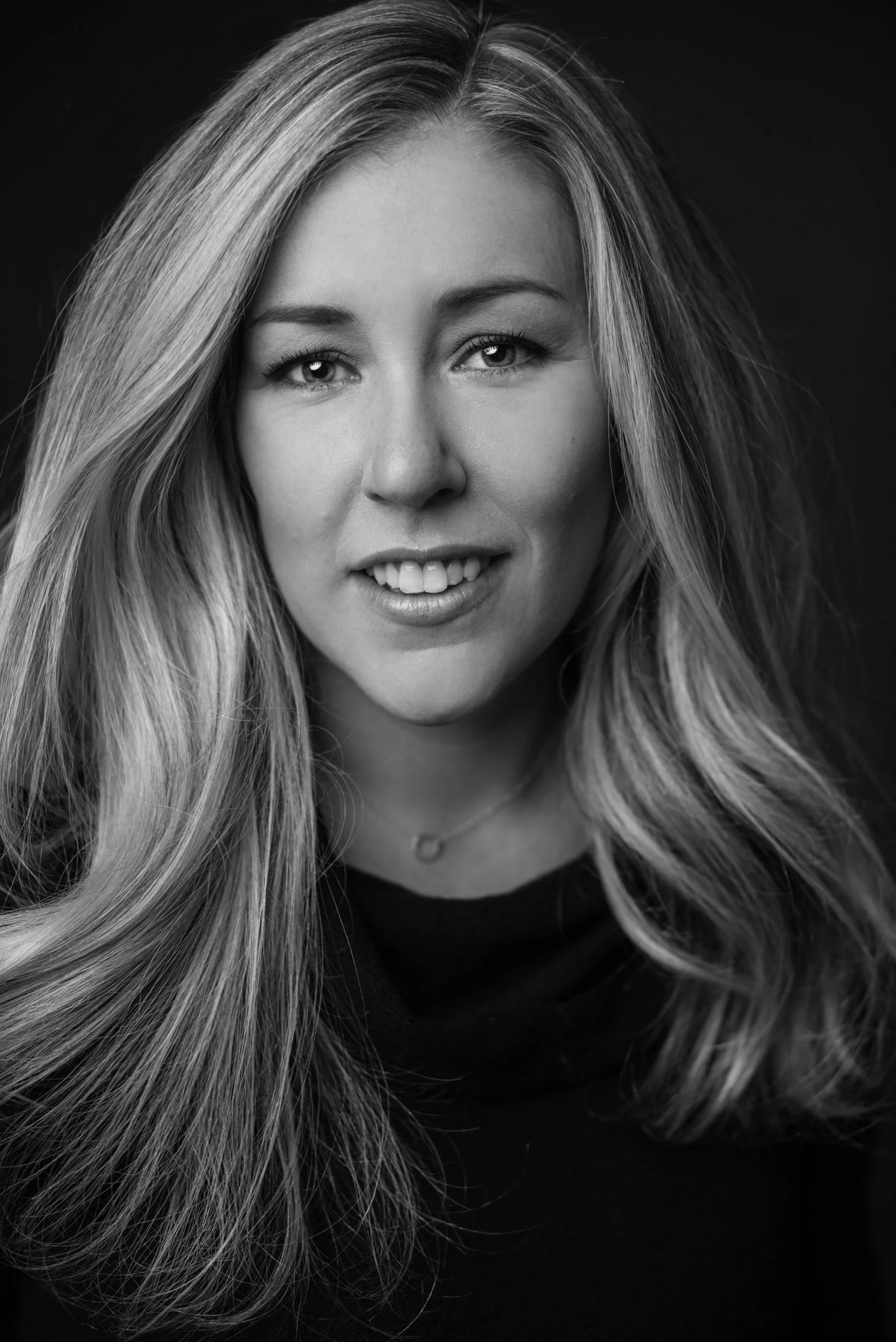 Black and white portrait of a woman with long, wavy hair, smiling slightly, wearing a dark top and a small heart-shaped necklace.