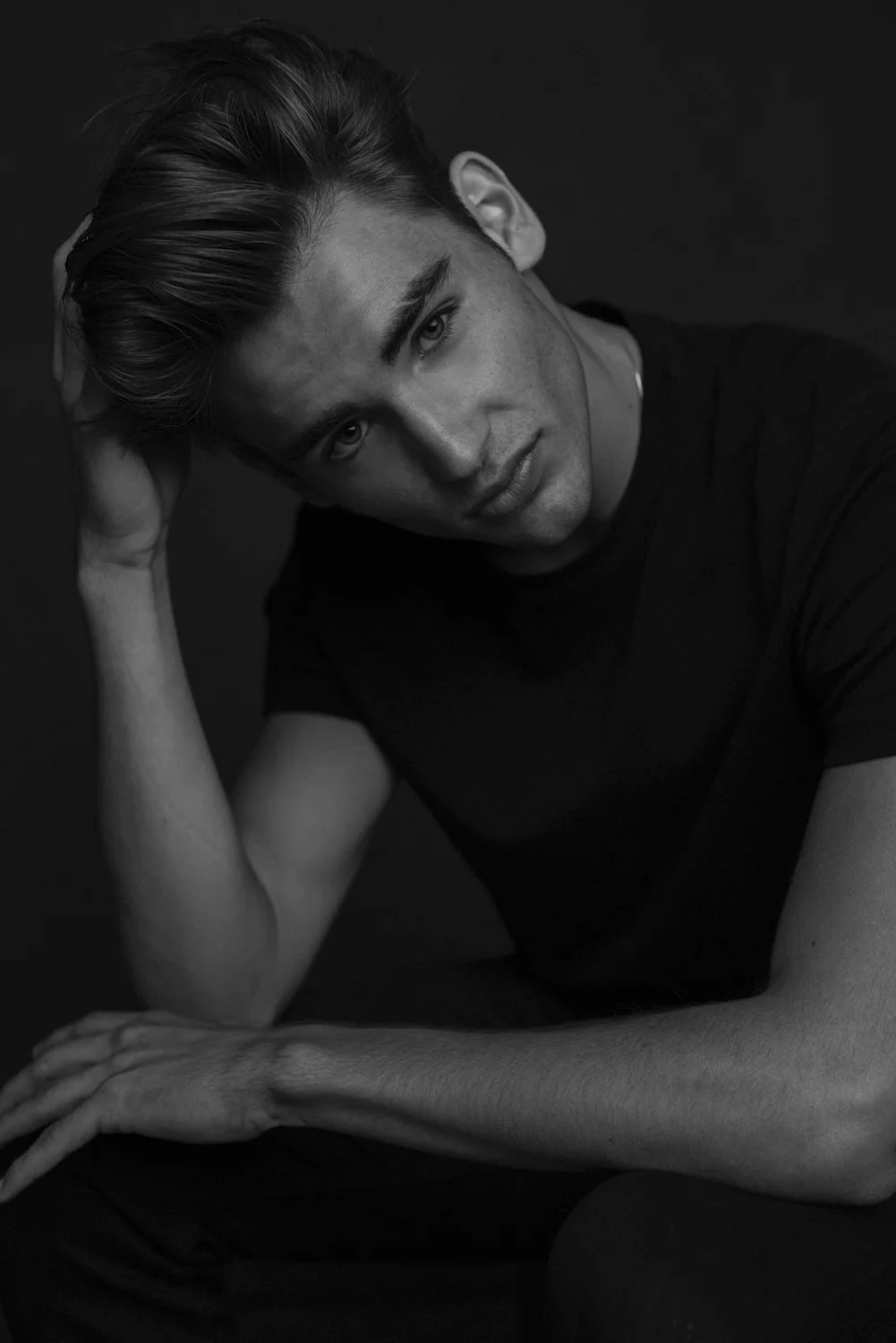 Black and white portrait of a young man with styled hair, wearing a dark t-shirt, resting his head on his right hand, with his left arm on his knee, looking directly at the camera against a dark background.