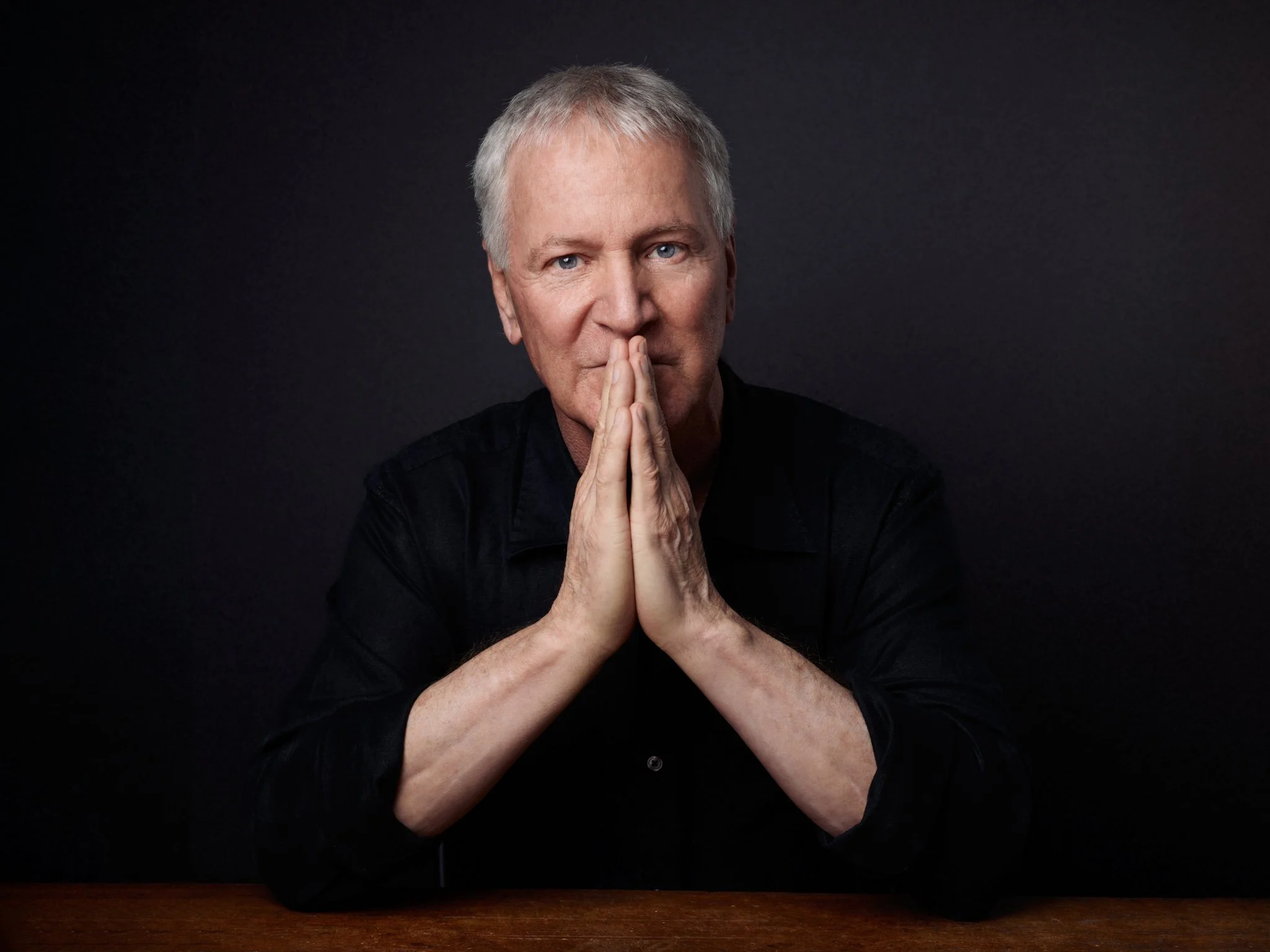 A middle-aged man with gray hair and blue eyes, wearing a black shirt, sitting at a wooden table with his hands pressed together near his face against a dark background.