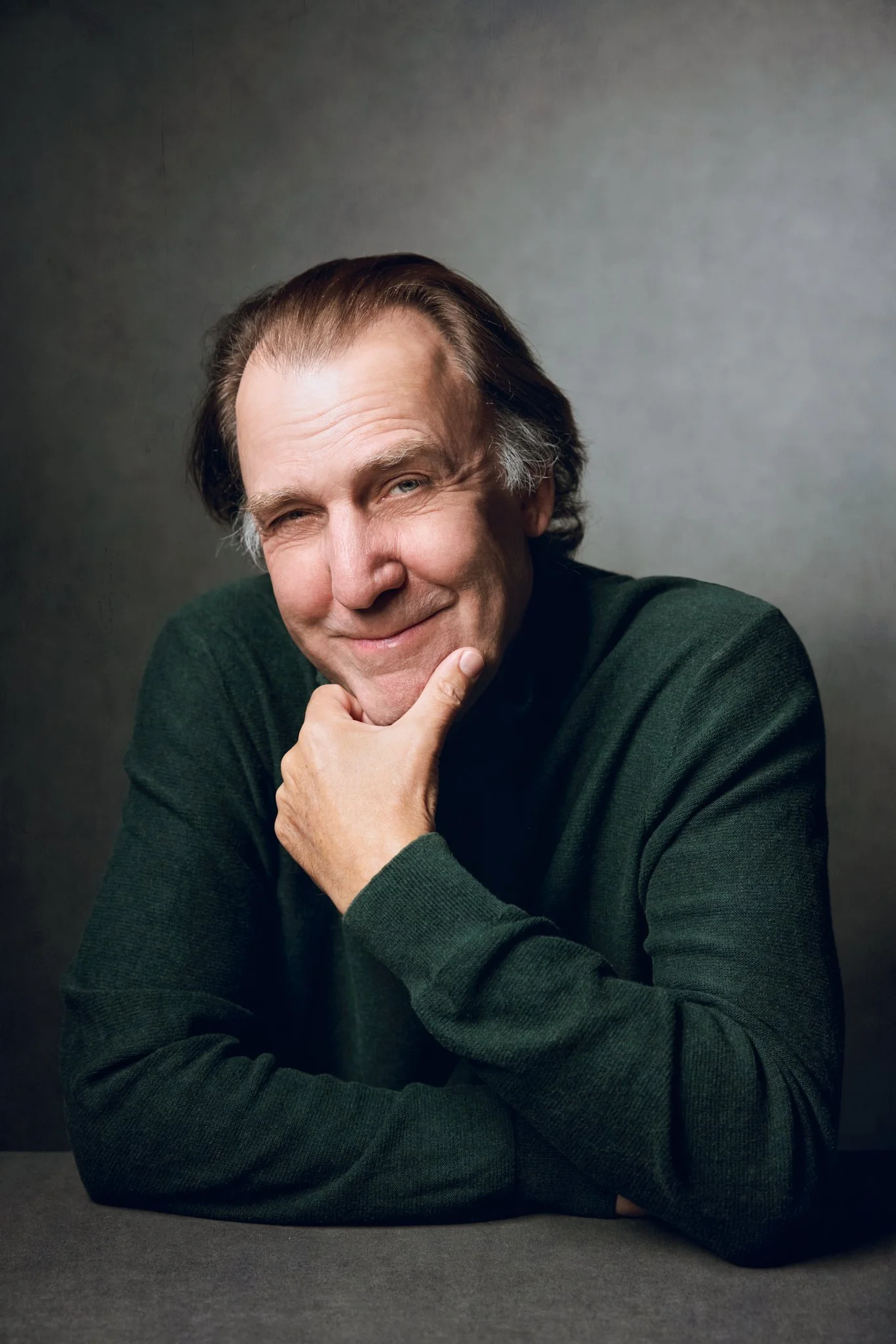 A middle-aged man with brown hair and a cheerful expression, sitting at a table with a gray background, wearing a dark green sweater.