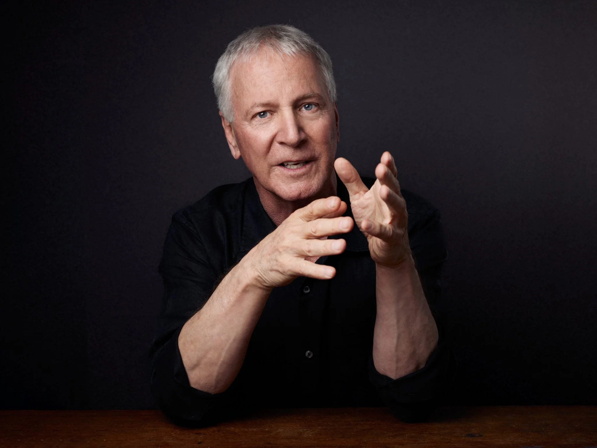 Older man with gray hair, wearing a black shirt, gesturing with his hands while speaking, against a dark background.