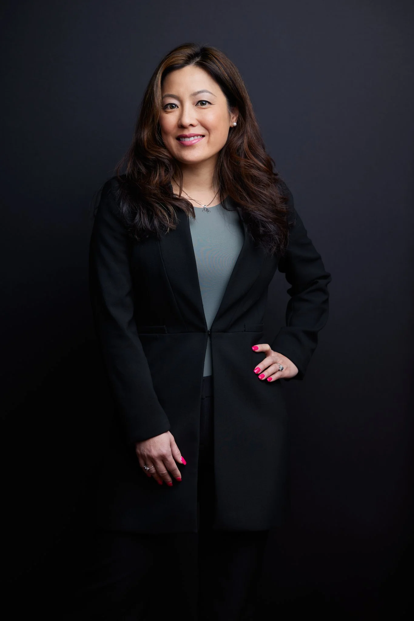 Professional woman with long wavy brown hair wearing a black blazer and gray top, smiling in front of a dark background.