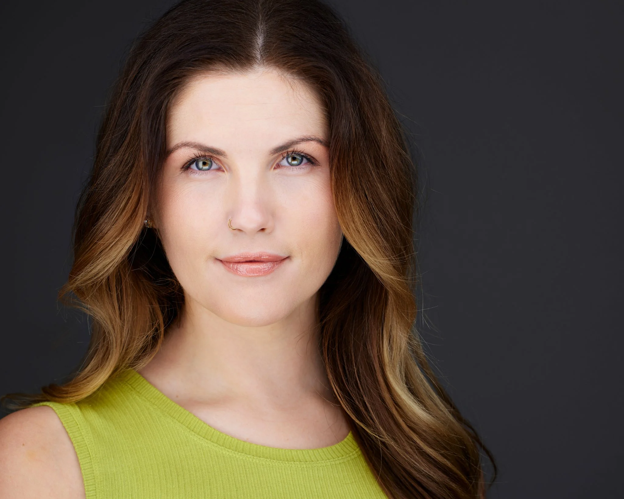Portrait of a woman with wavy brown hair, blue eyes, wearing a lime green sleeveless top, against a dark gray background.