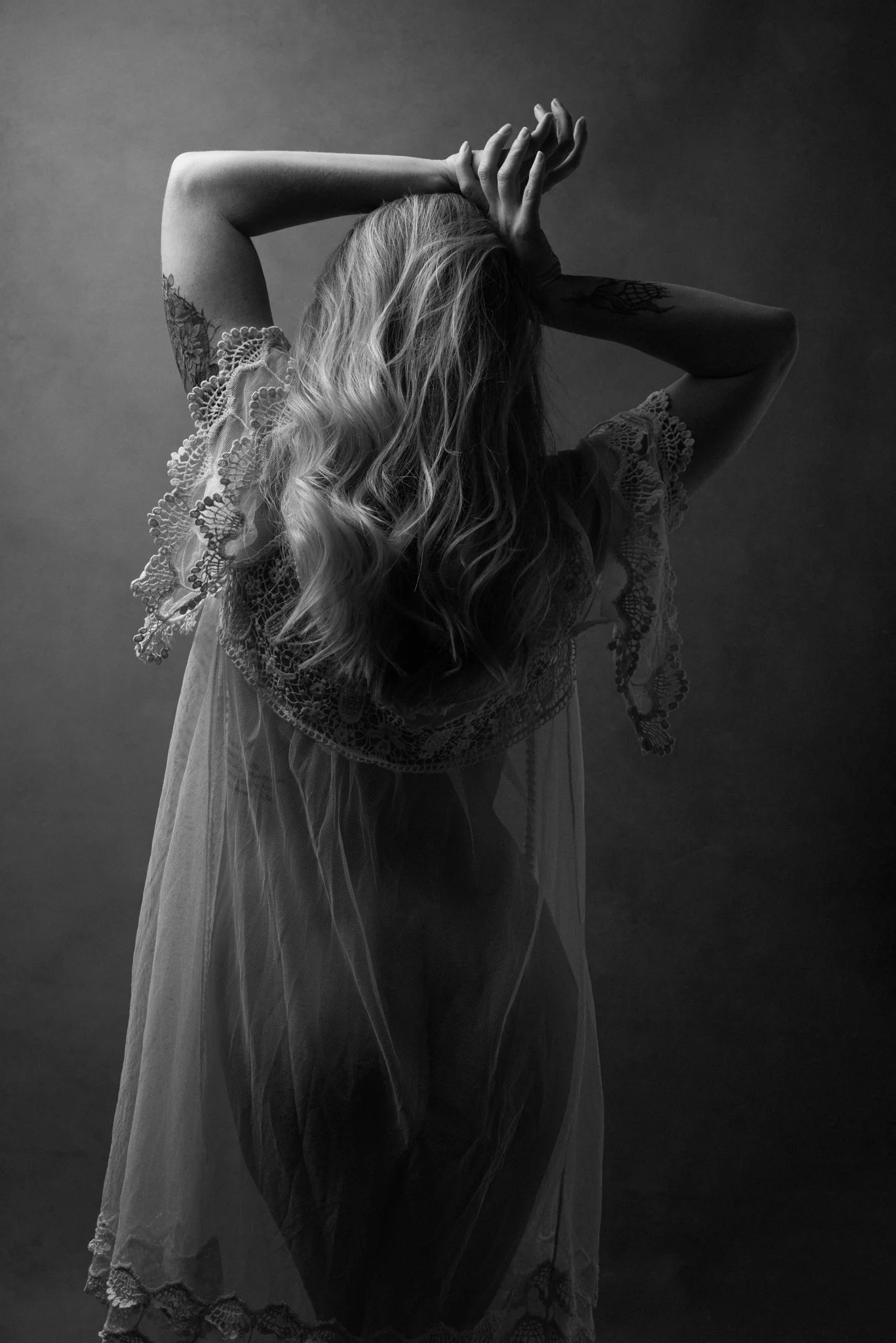 A woman with long, wavy hair in a lace dress poses with her arms raised above her head, obscuring her face, in a black and white photo.