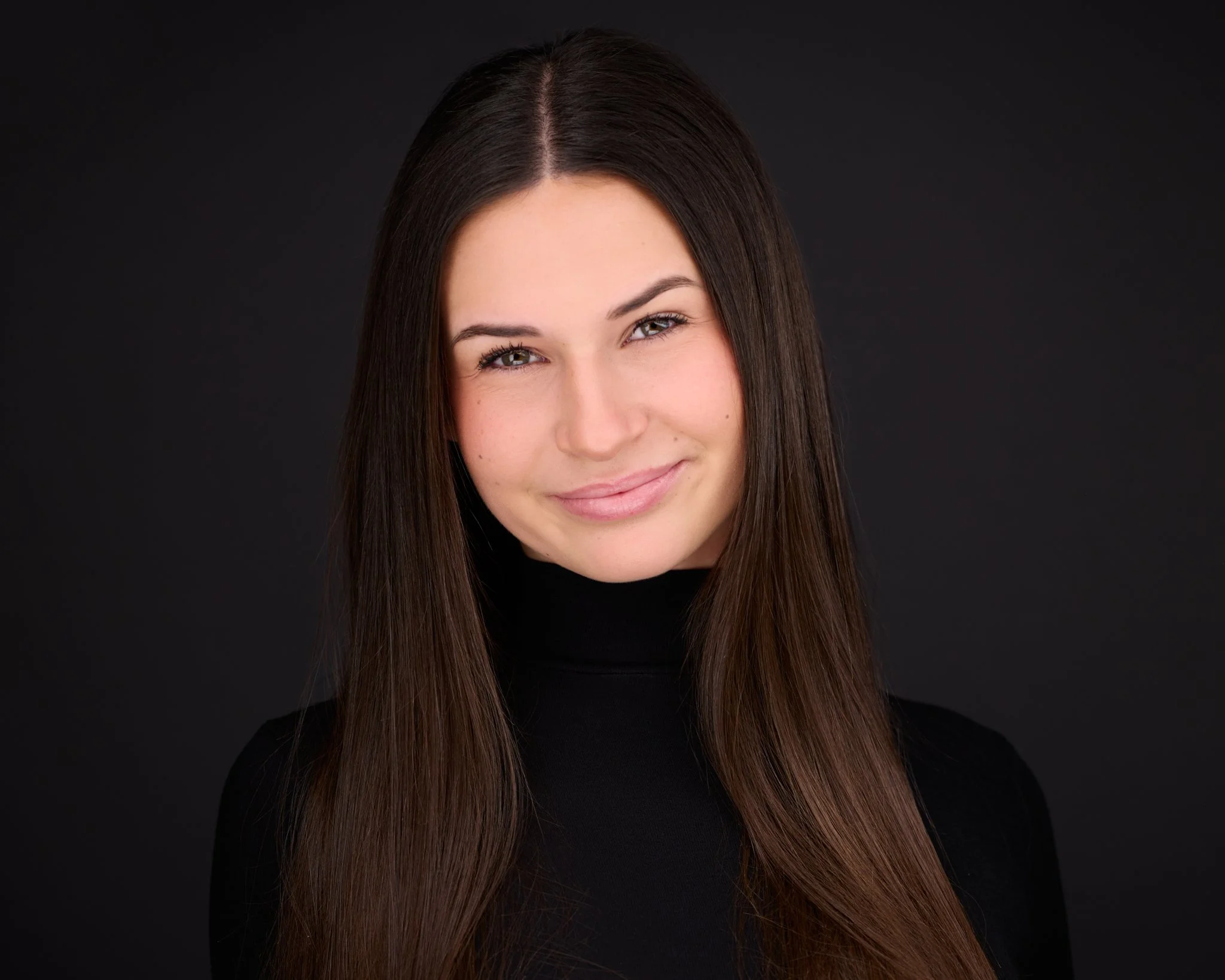 A young woman with long brown hair wearing a black top, smiling softly against a dark background.