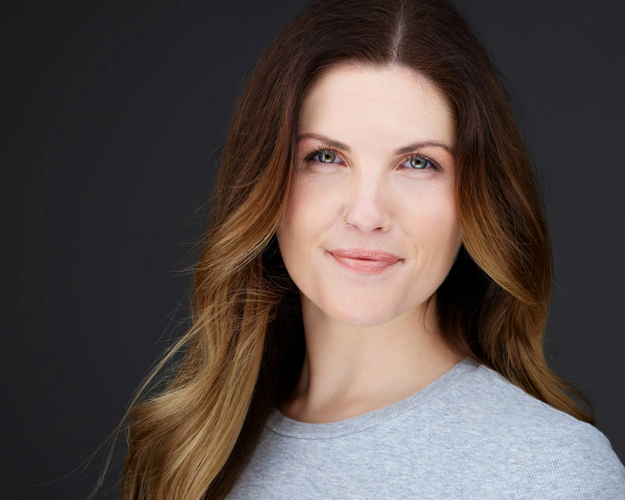A woman with long, wavy, light brown hair, wearing a light gray top, smiling softly at the camera against a dark background.